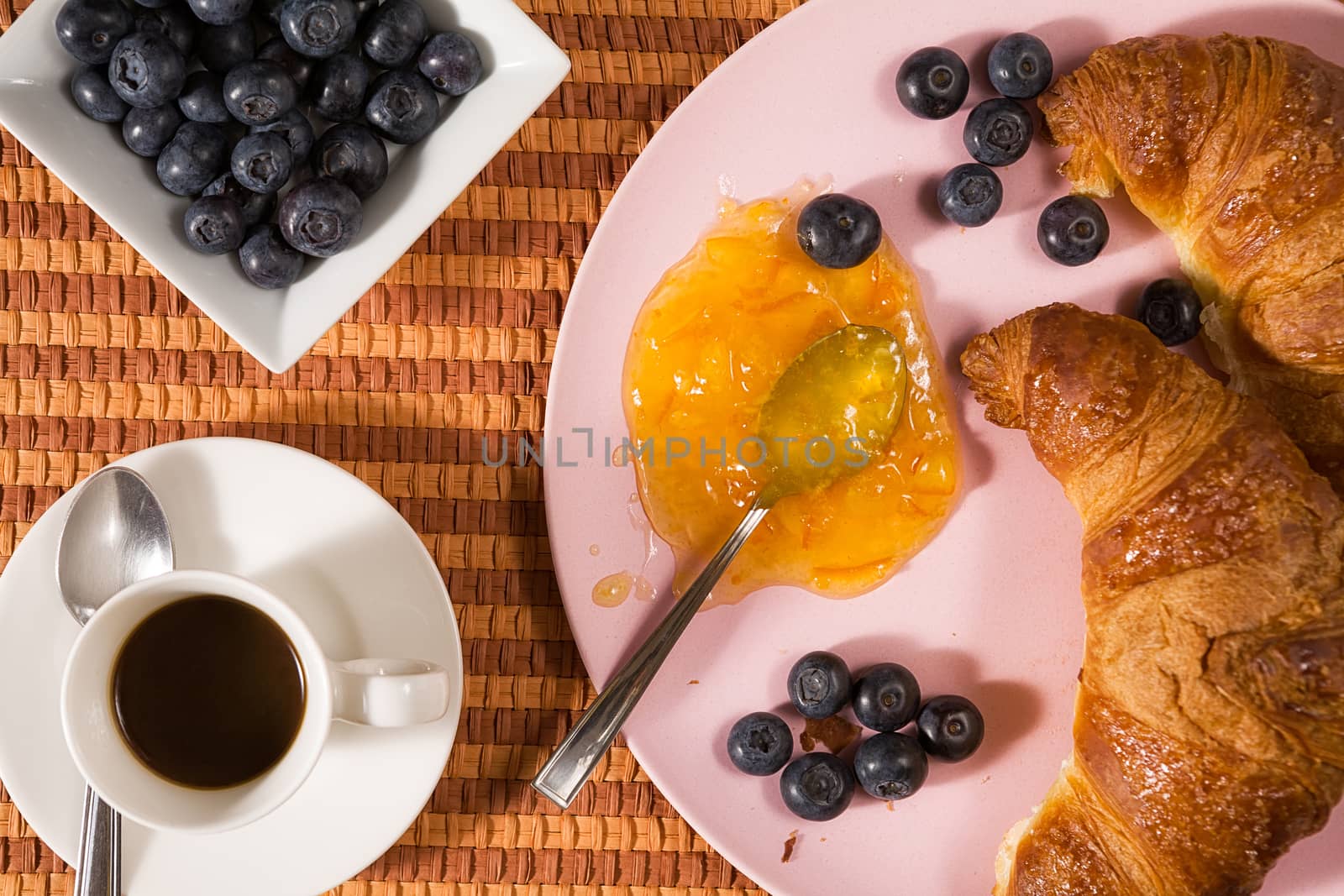 Closeup of croissant, blueberries, coffee and orange jam over a tablecloth seen from above