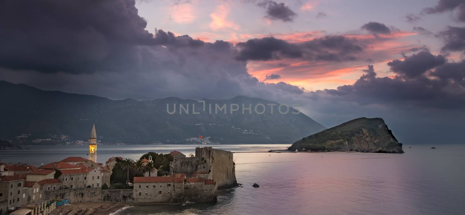 Sunrise over Budva old town with lit tower of Sv. Ivan church, old town's citadel and island Sv. Nikola to the right. Picturesque mountains and coast in background