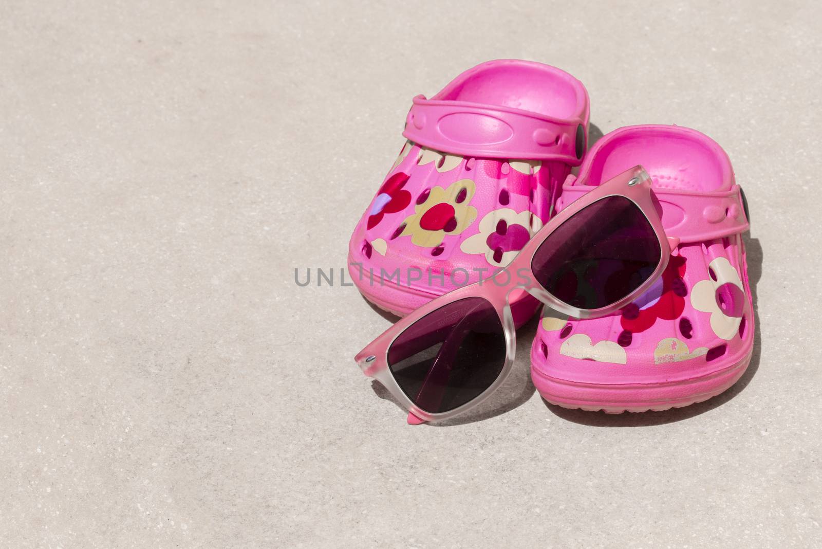 Pink beach kid's crocs and sunglasses on sandy beach.Beach sandals and sunglasses in the foreground and blurred sea in the background.