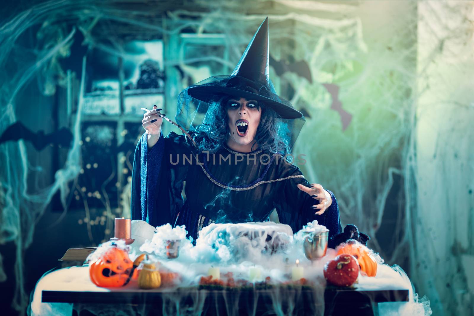 Witch with awfully face and hat on her head in creepy surroundings full of cobweb sends evil. Halloween concept.