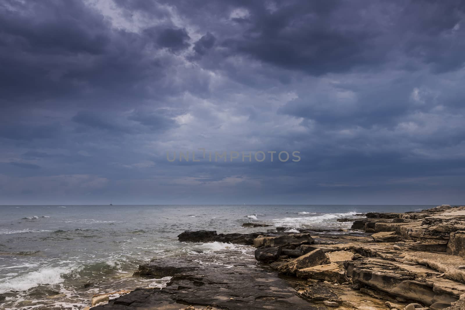 Dramatic skies over a rocky beach and sail boats in distance on horizon