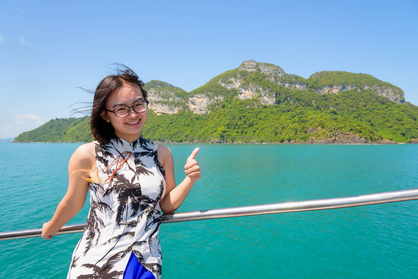 Cute young woman with eyeglasses thumb up the beautiful natural of the sea island and sky in summer on the boat while cruising at Mu Ko Ang Thong National Park, Surat Thani, Thailand