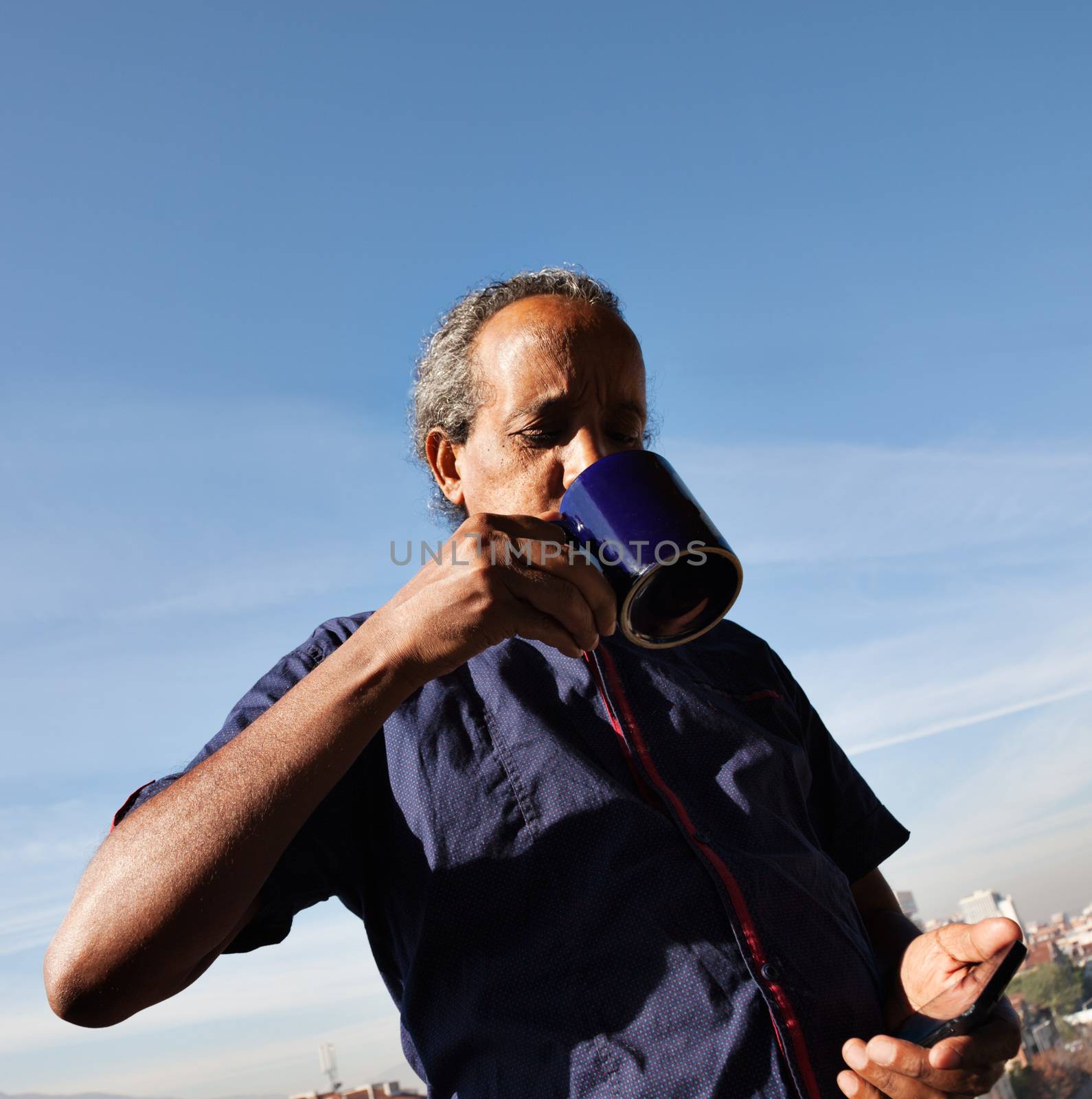 Rasta black man drinking cofee or tea, holding phone, blue sky behind.