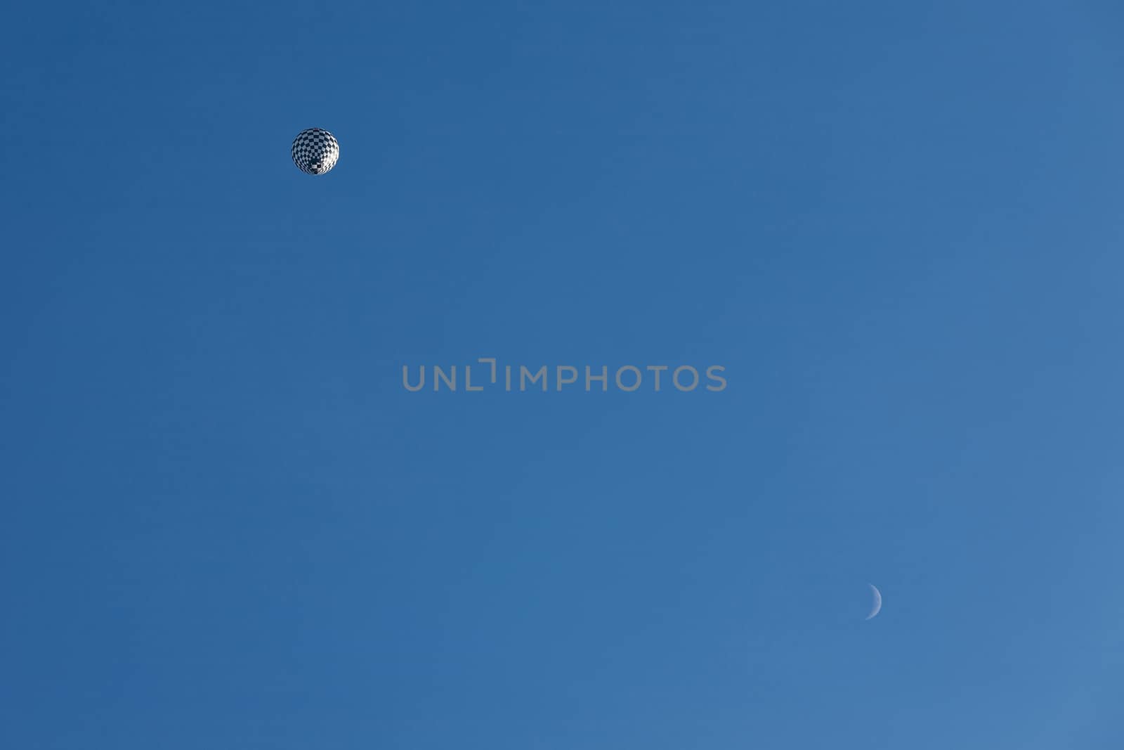 Black and white balloon in flight and moon seen from below against a blue sky