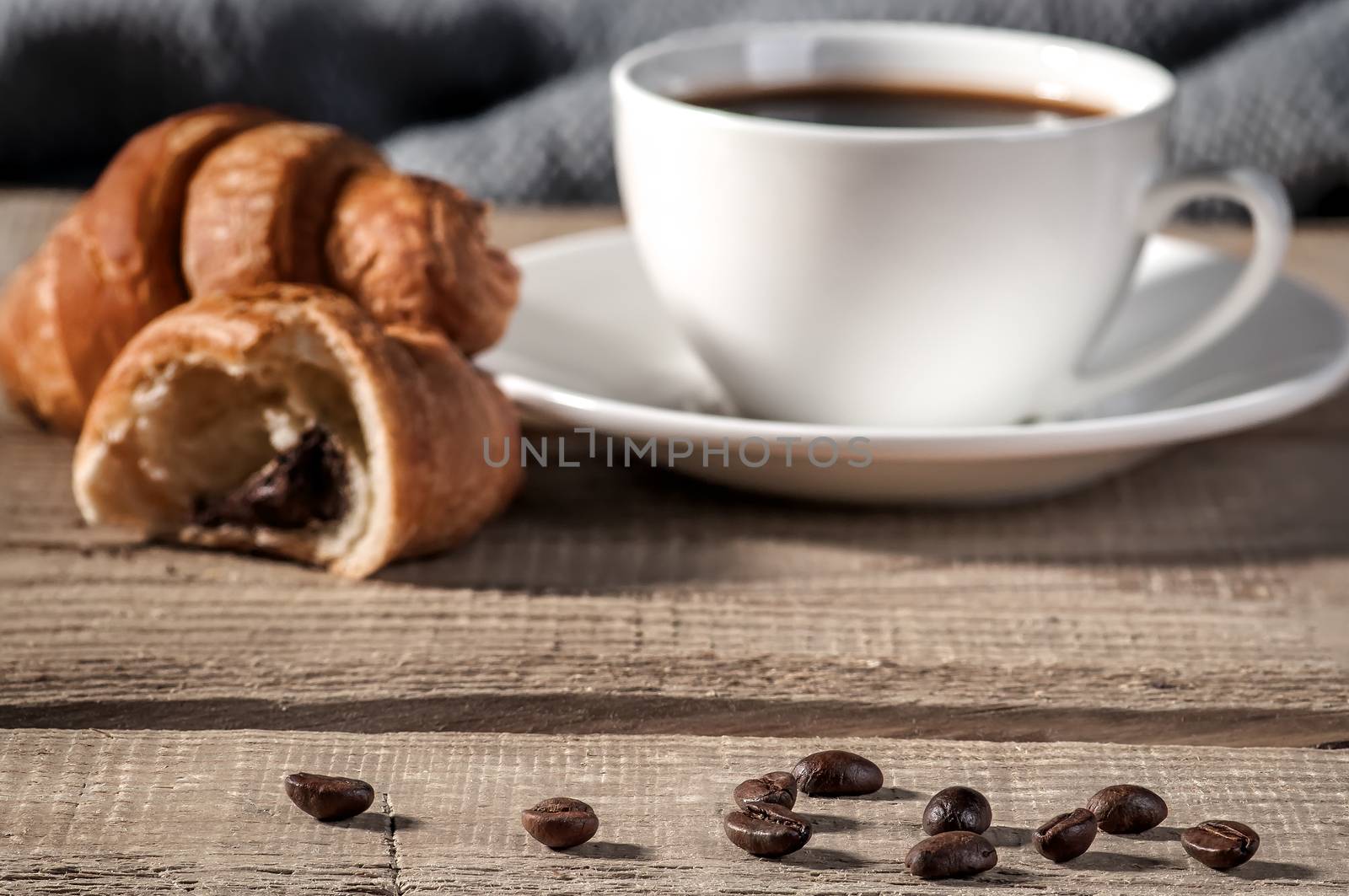 Coffee beans on the wooden table, blurred cup of coffee and a croissant in the background