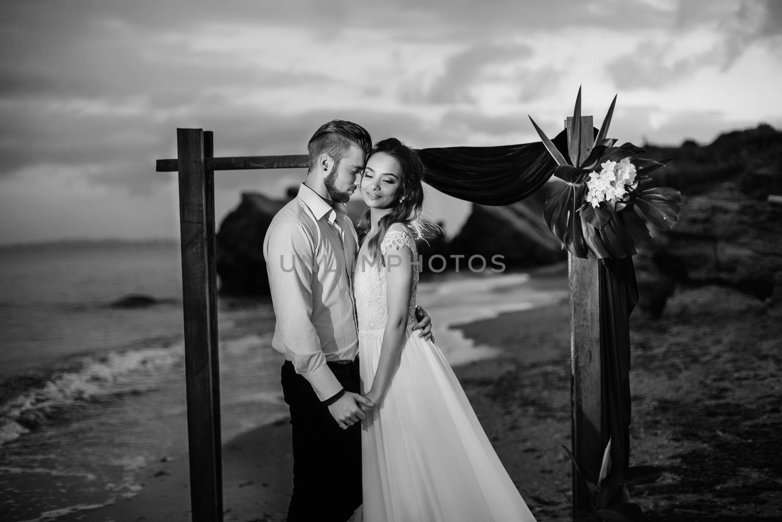 young couple groom and bride with a bouquet in the evening on the beach near the wedding arch