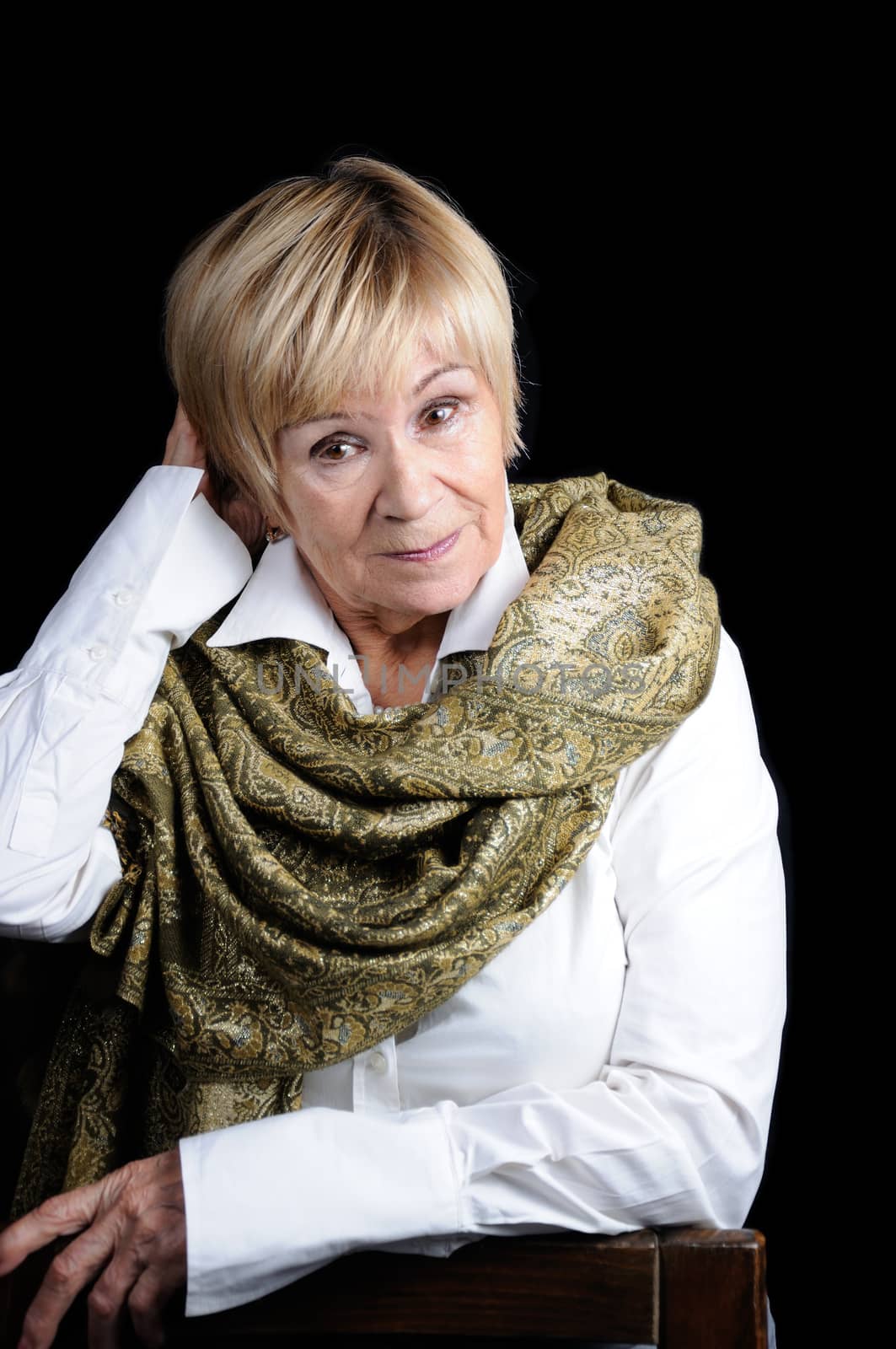 Portrait of an elderly woman adjusting her hair in a white shirt with a shawl on her shoulders against a dark background close-up