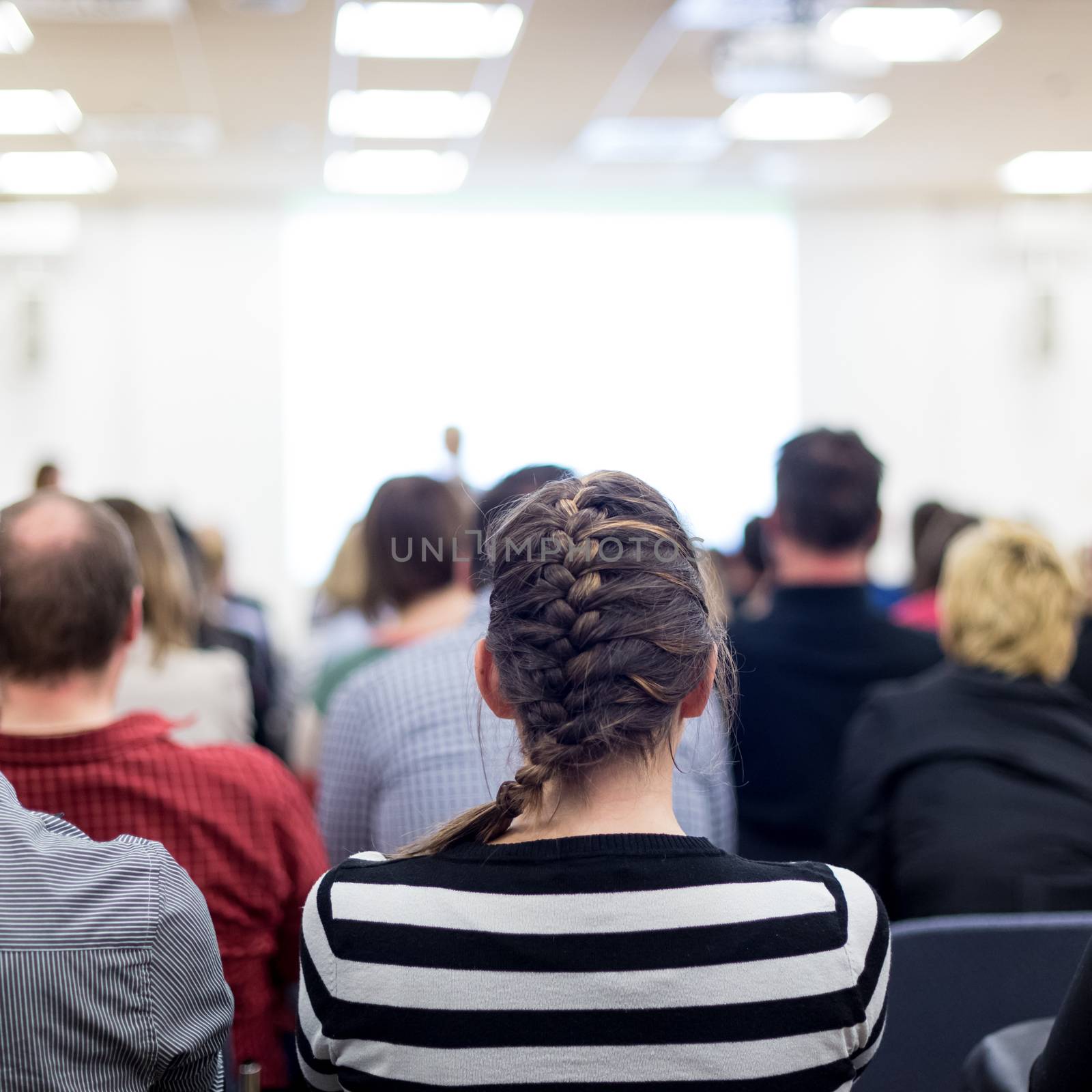 Business and entrepreneurship symposium. Female speaker giving a talk at business meeting. Audience in conference hall. Rear view of unrecognized participant in audience. Copy space on whitescreen.