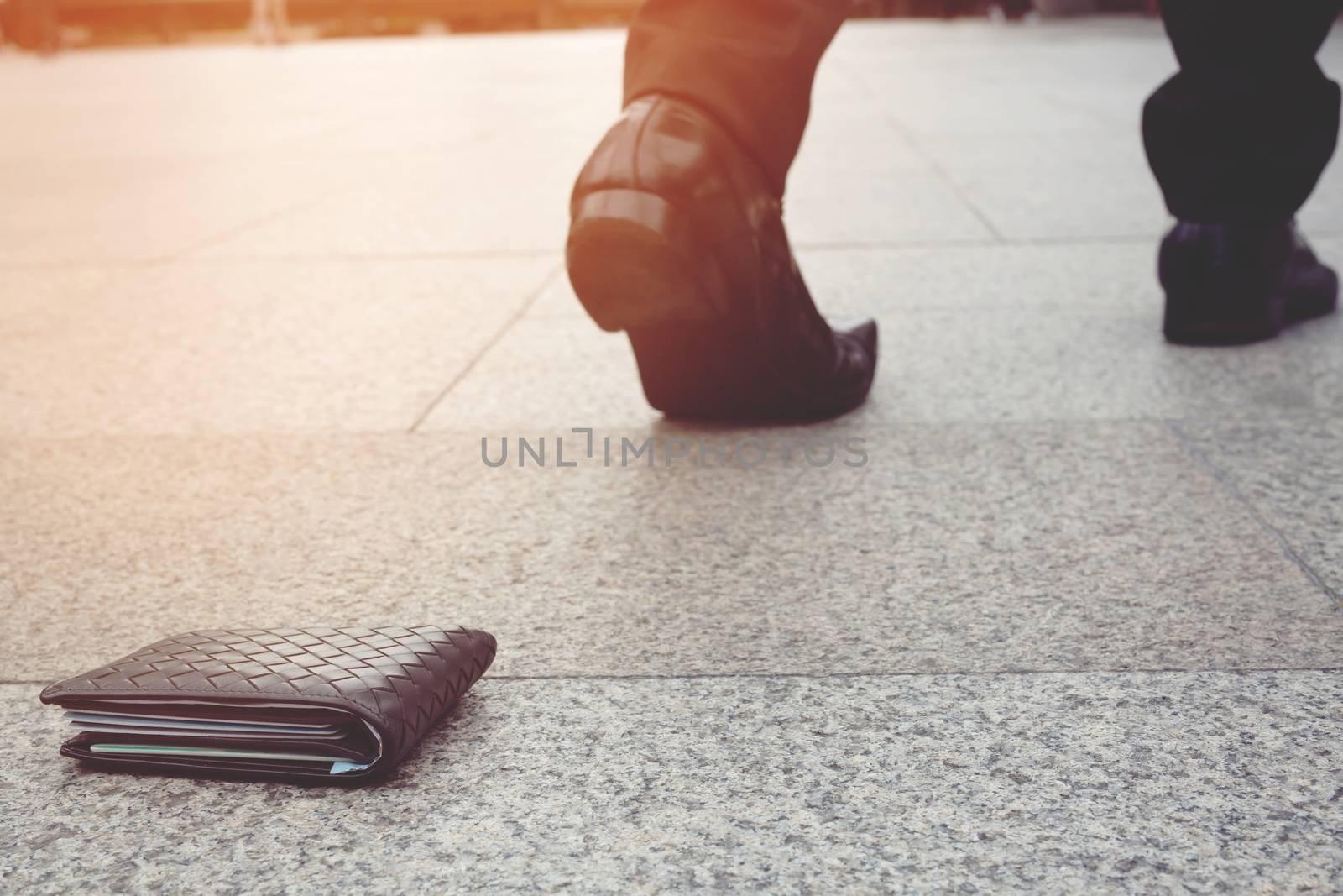 Close-up shot of brown leather wallet laying at sidewalk, and feet of woman going away on the background. Concept of money loss