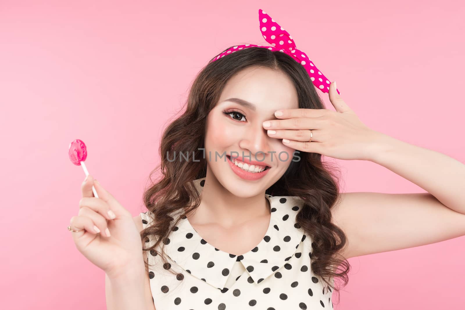 Smiling cute girl with lollipop, covering her eye by hand over pink background