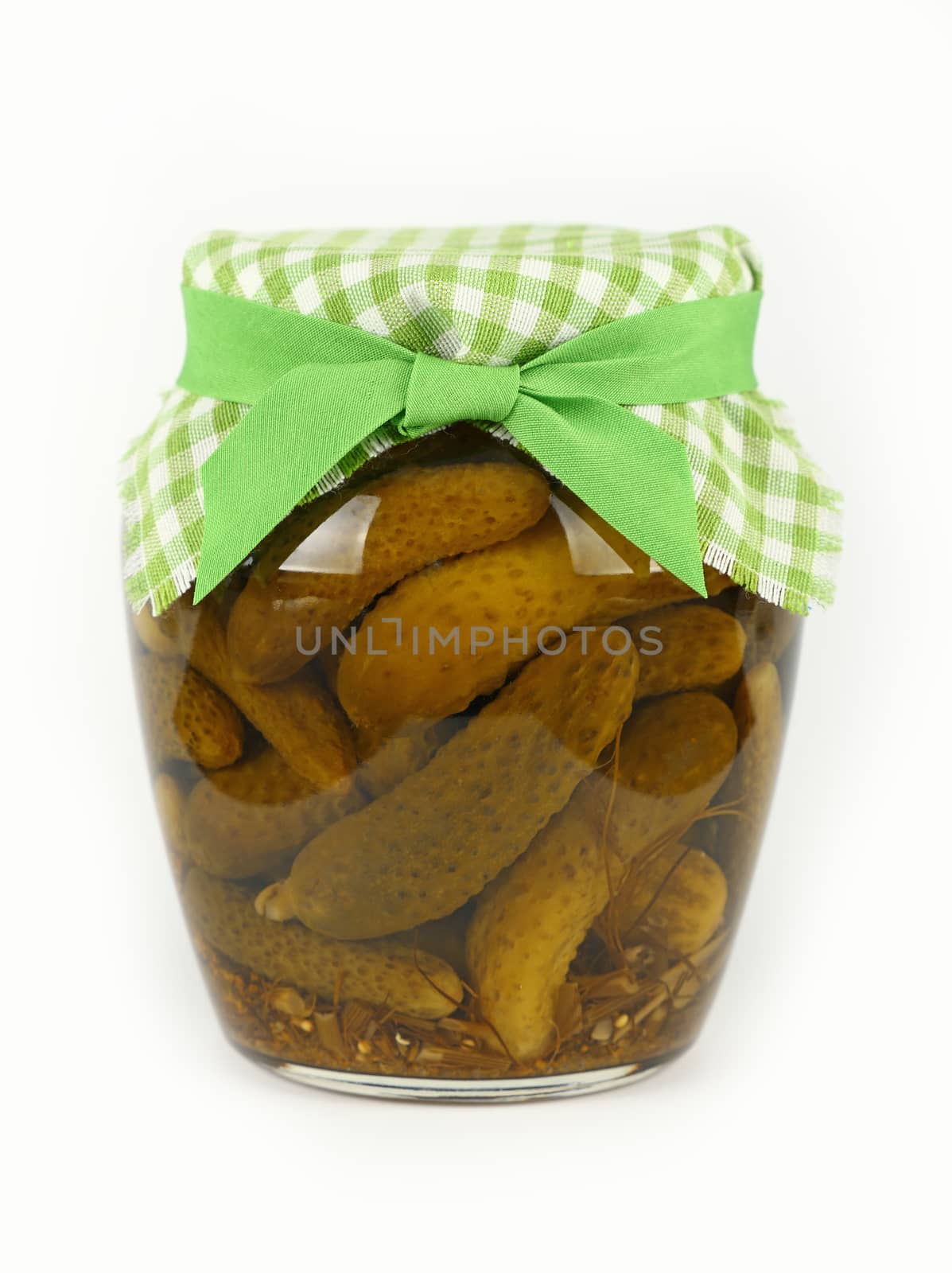 Close up of one glass jar of pickled small gherkin cucumbers checkered linen textile fabric lid decoration and green ribbon over white background, low angle side view