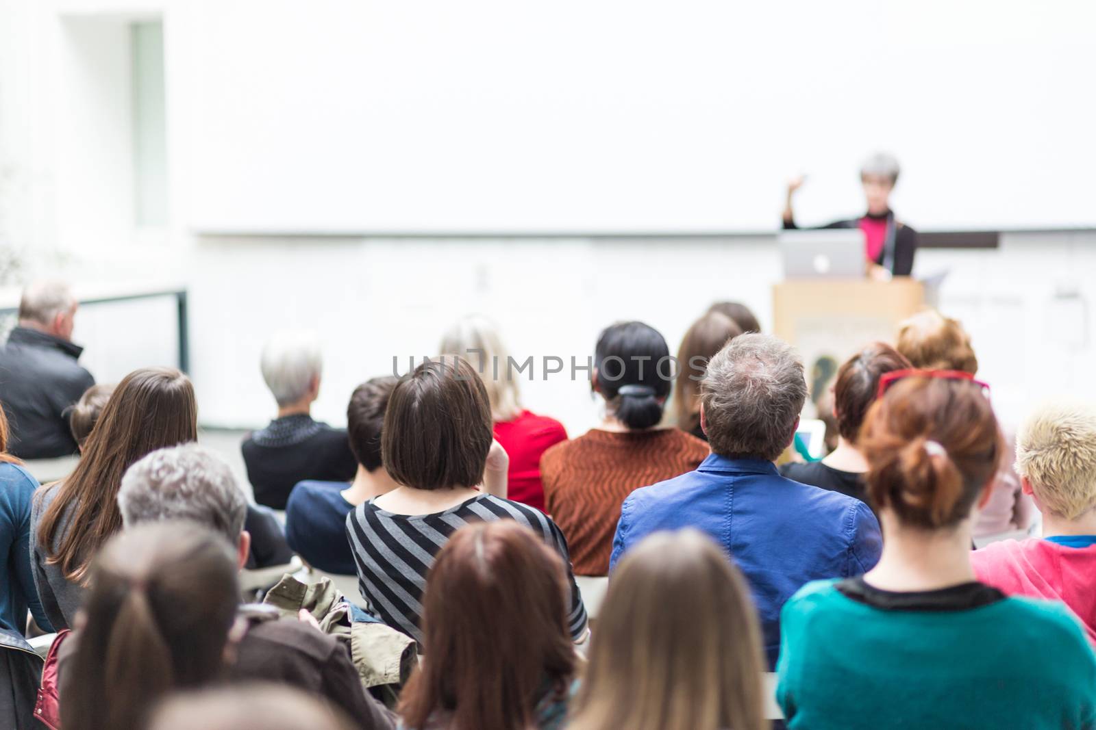 Business and entrepreneurship symposium. Female speaker giving a talk at business meeting. Audience in conference hall. Rear view of unrecognized participant in audience.