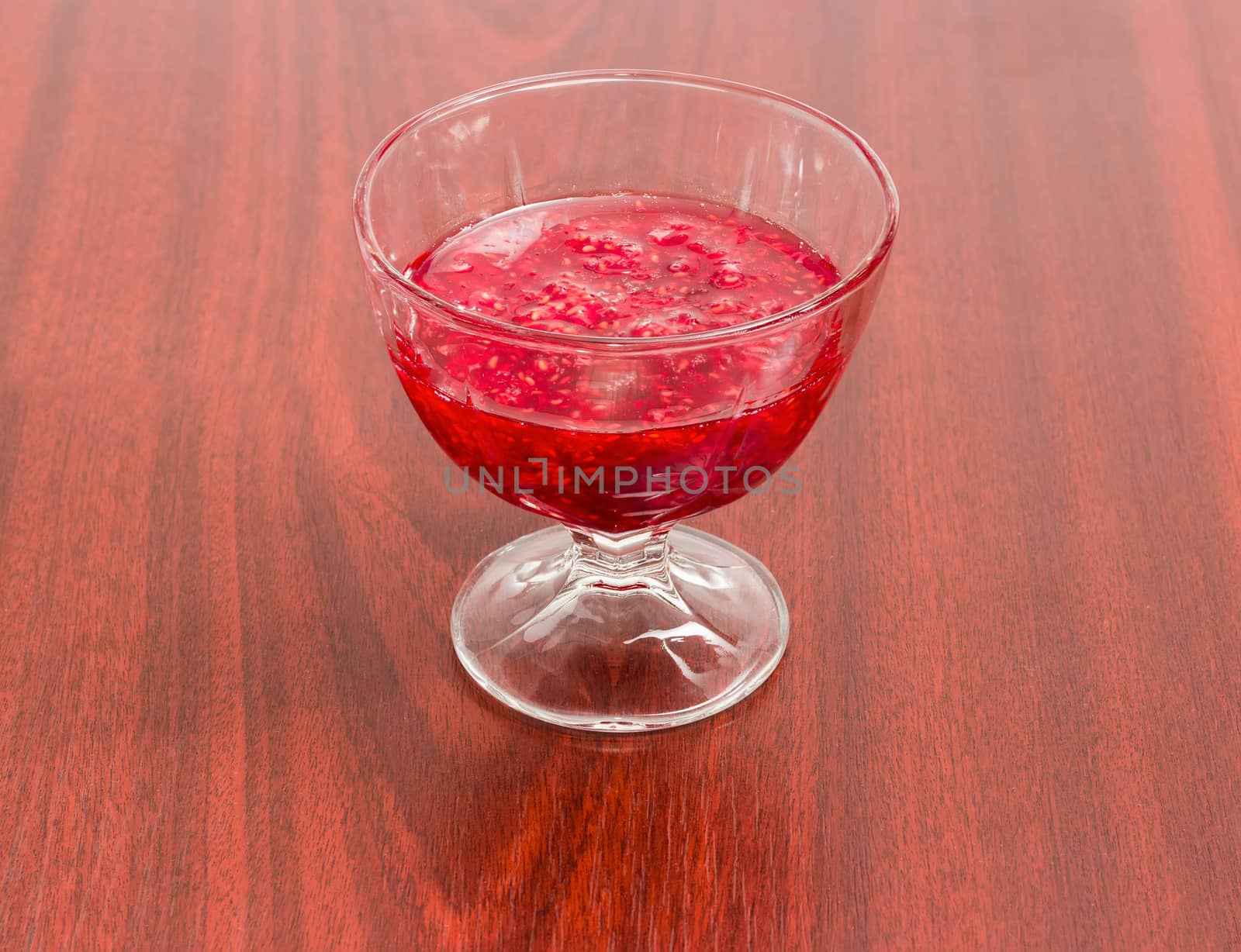 Raspberry jam in the glass dessert stem bowl on a red wooden table
