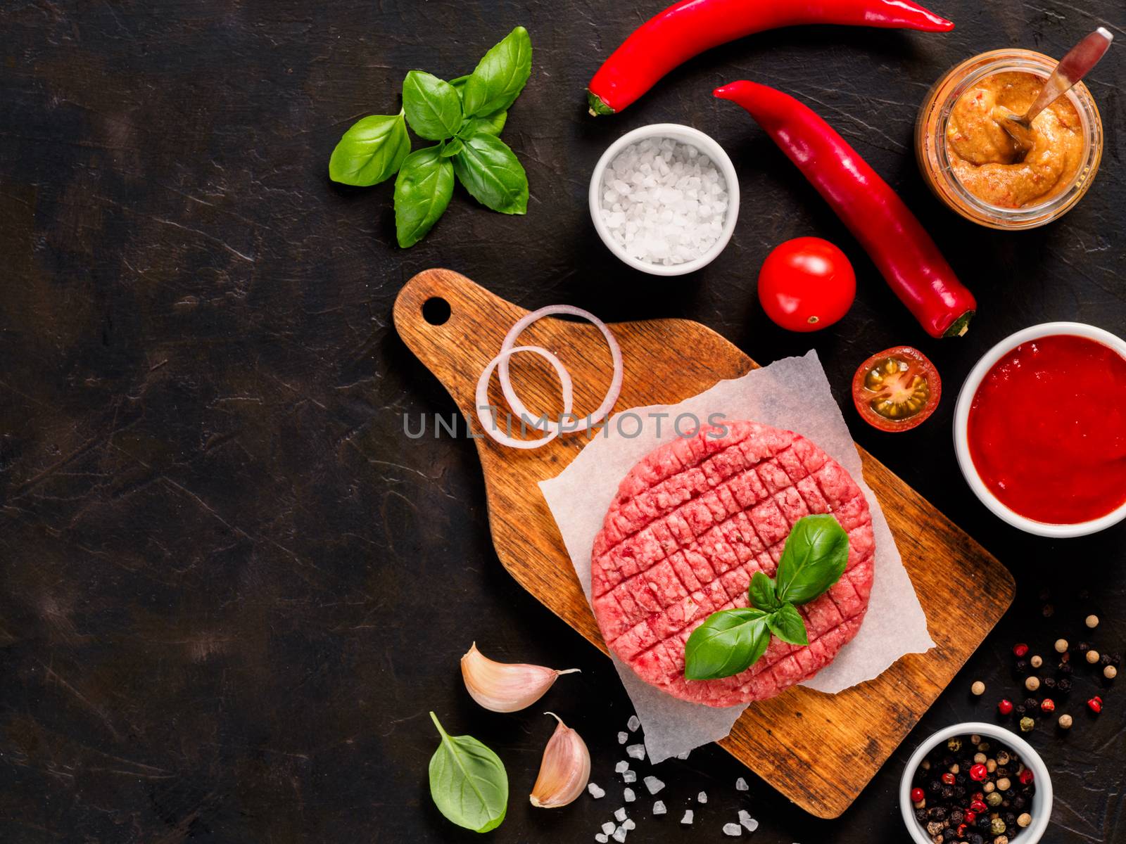 Raw beef meat steak cutlet for burger on wooden cutting board with spices, vegetables, sauces ketchup and mustard on black concrete background. Making homemade burger. Top view or flat lay. Copy space