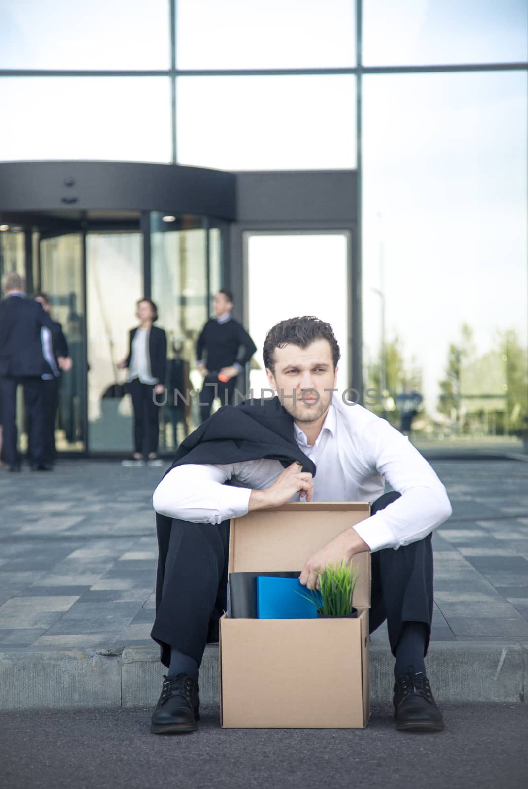 Fired business man sitting frustrated and upset on the street near office building with box of his belongings. He lost work