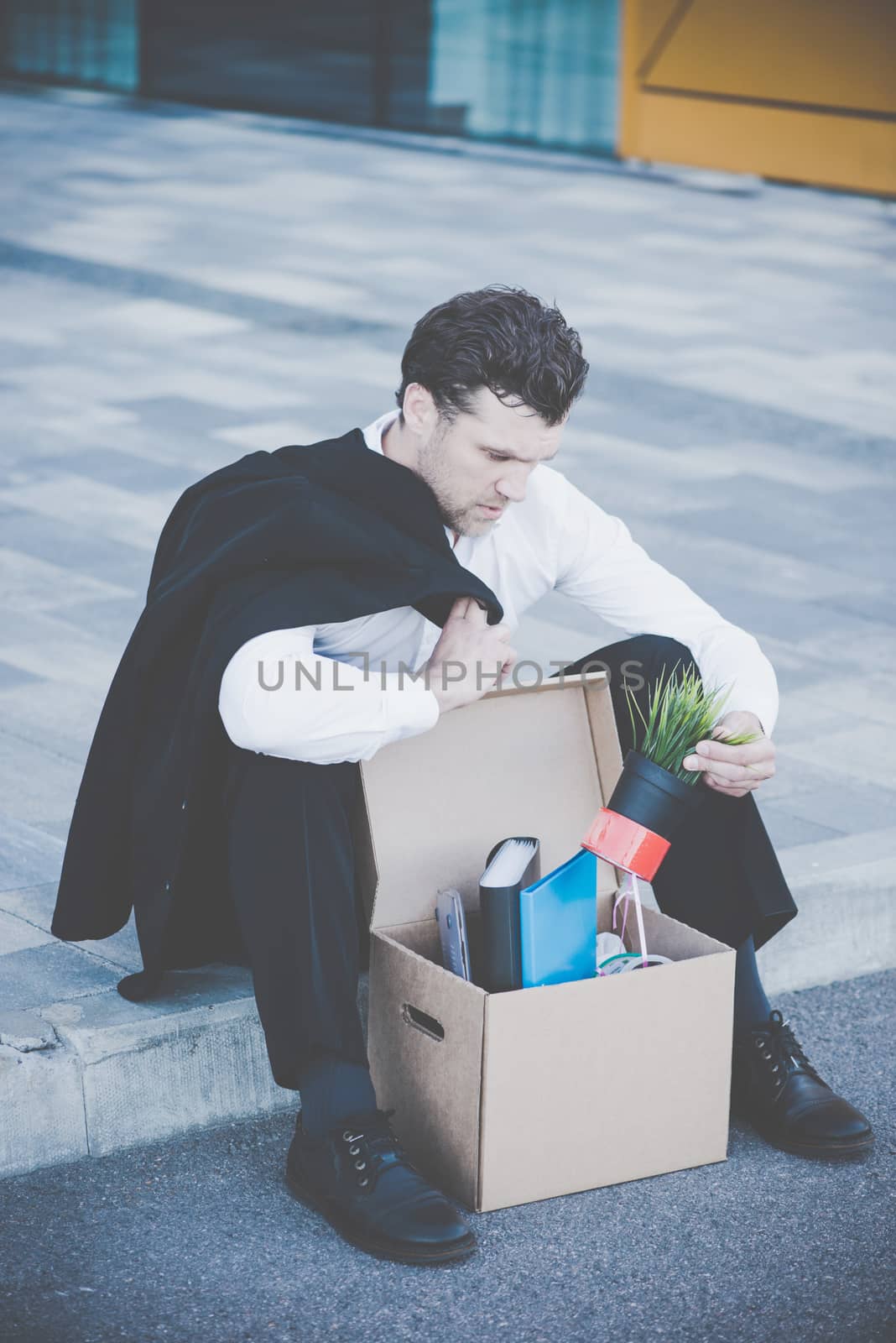 Fired business man sitting frustrated and upset on the street near office building with box of his belongings. He lost work