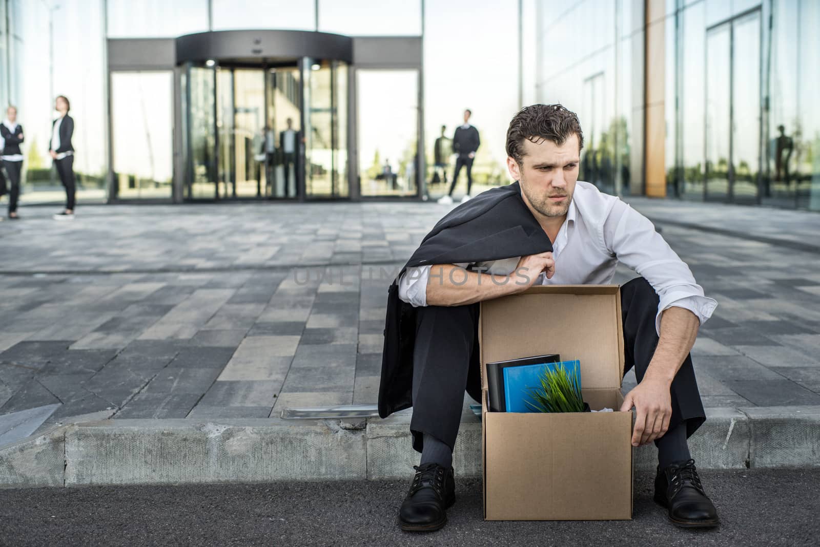 Fired business man sitting frustrated and upset on the street near office building with box of his belongings. He lost work