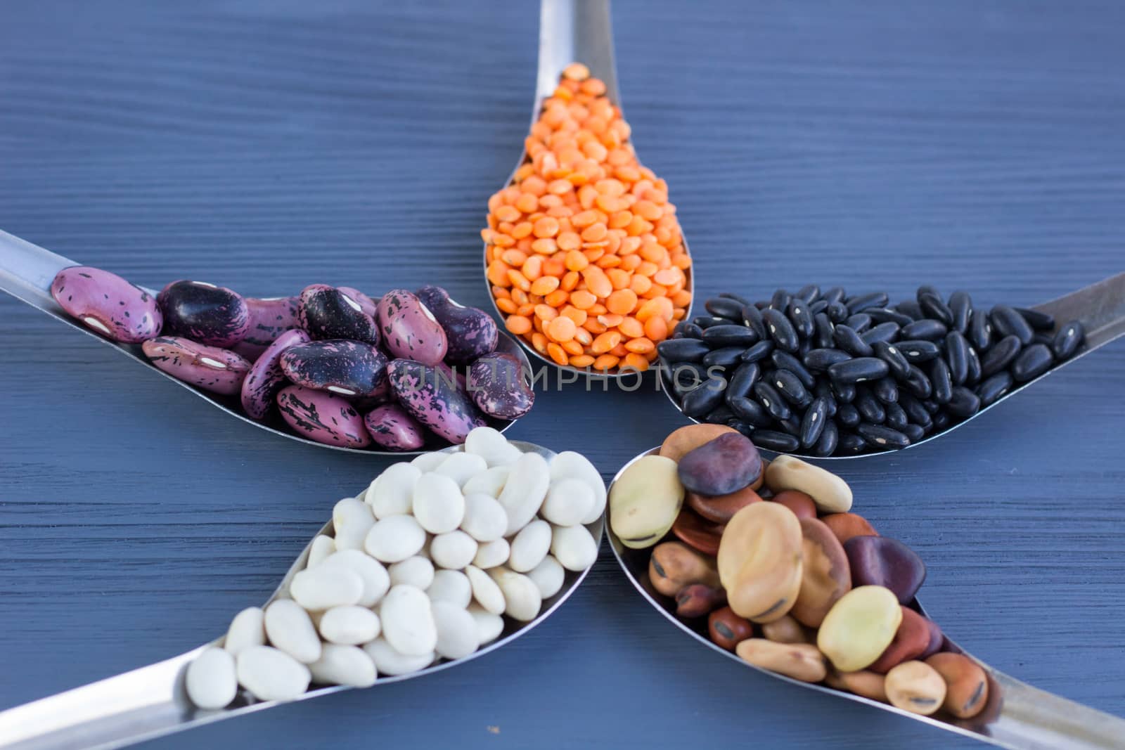 Yellow lentils, black, white, brown, purple kidney beans on metal spoons star composition on blue wooden background