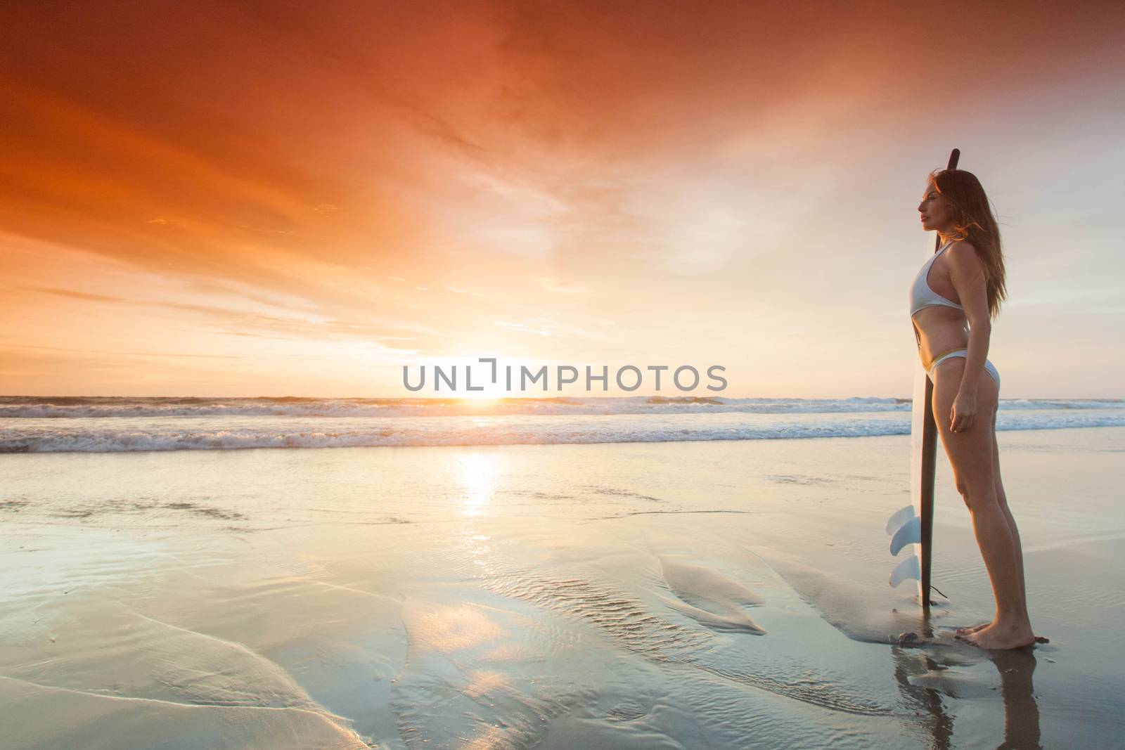 A beautiful young slim sporty woman in bikini with a surfboard is standing at ocean beach at sunset