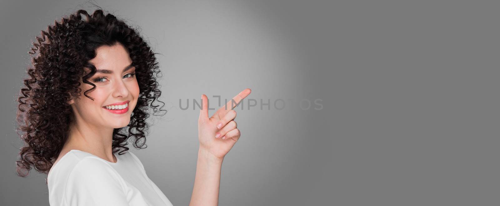 Portrait of amazing beautiful smiling woman with curly hair on gray background pointing at copy space