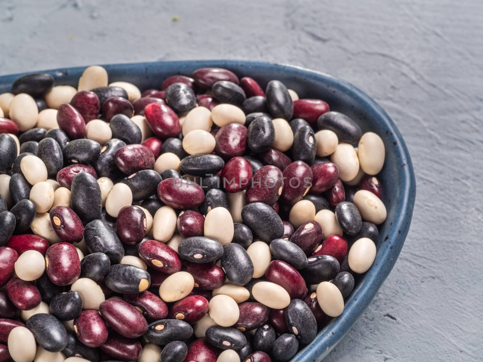 Close up view of raw mixed beans in blue plate on gray concrete background. Mixed of black, red and white beans with copy space.