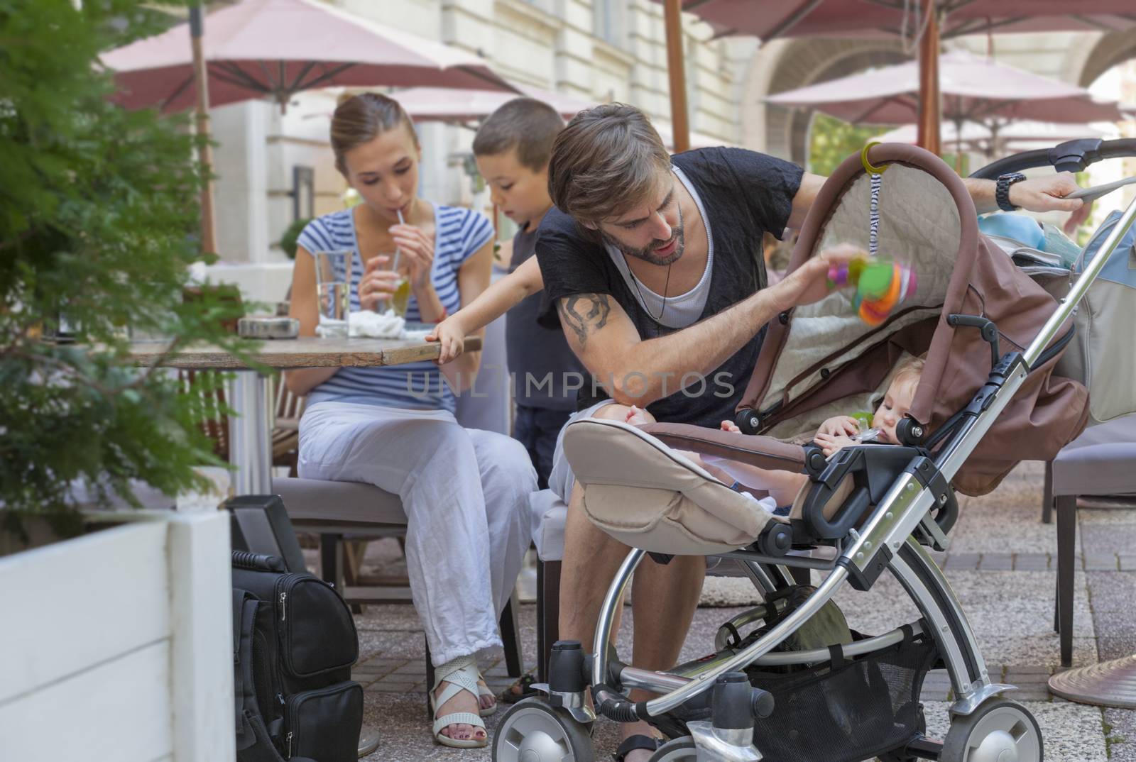 A father is taking care of his baby, while her life and other young boy are communicating in background. Cafe outdoors, summer.