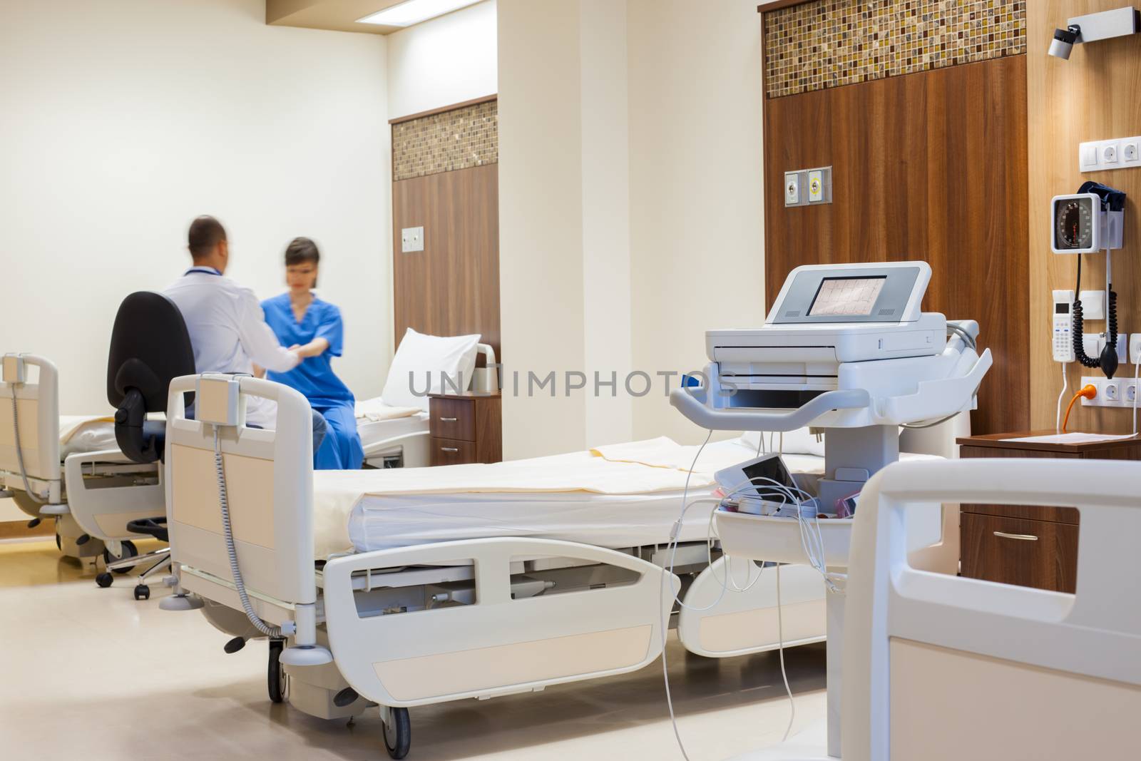 Blurred figure of a patient being helped by a doctor to rise from the bed in a spacy modern hospital room. ECG machine on focus.