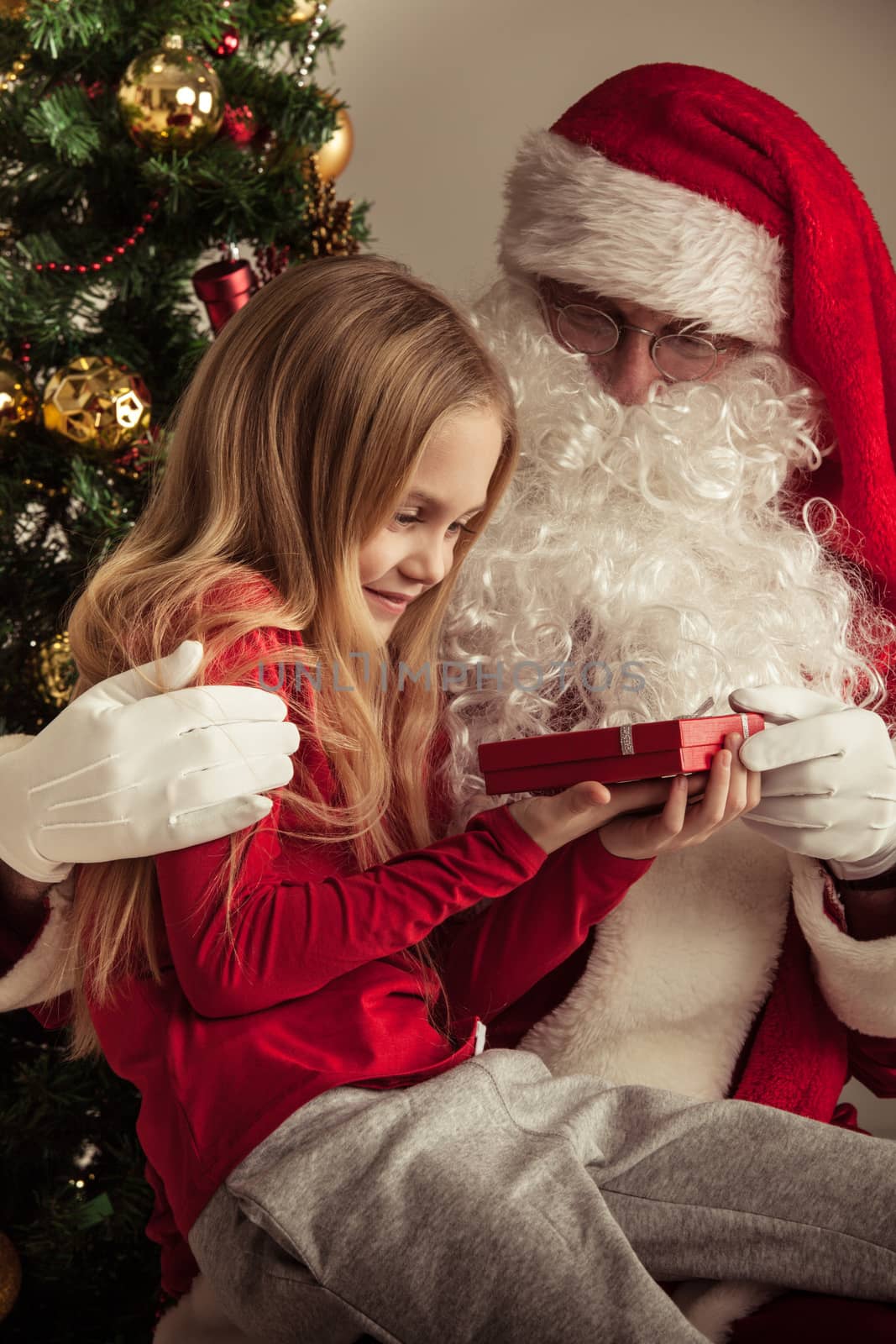 Portrait of smiling little girl sitting on santa claus knees near christmas tree and getting a gift