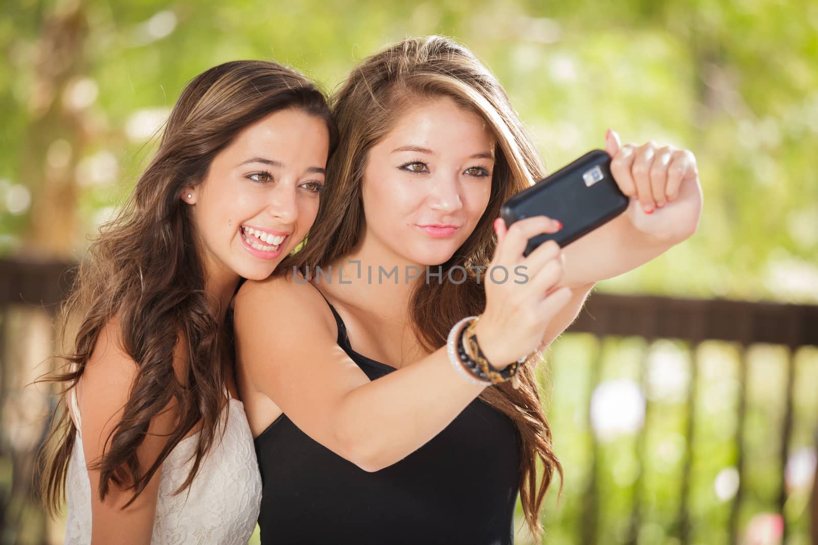 Two Attractive Mixed Race Girlfriends Using Their Smart Cell Phones For Selfie Outdoors by Feverpitched