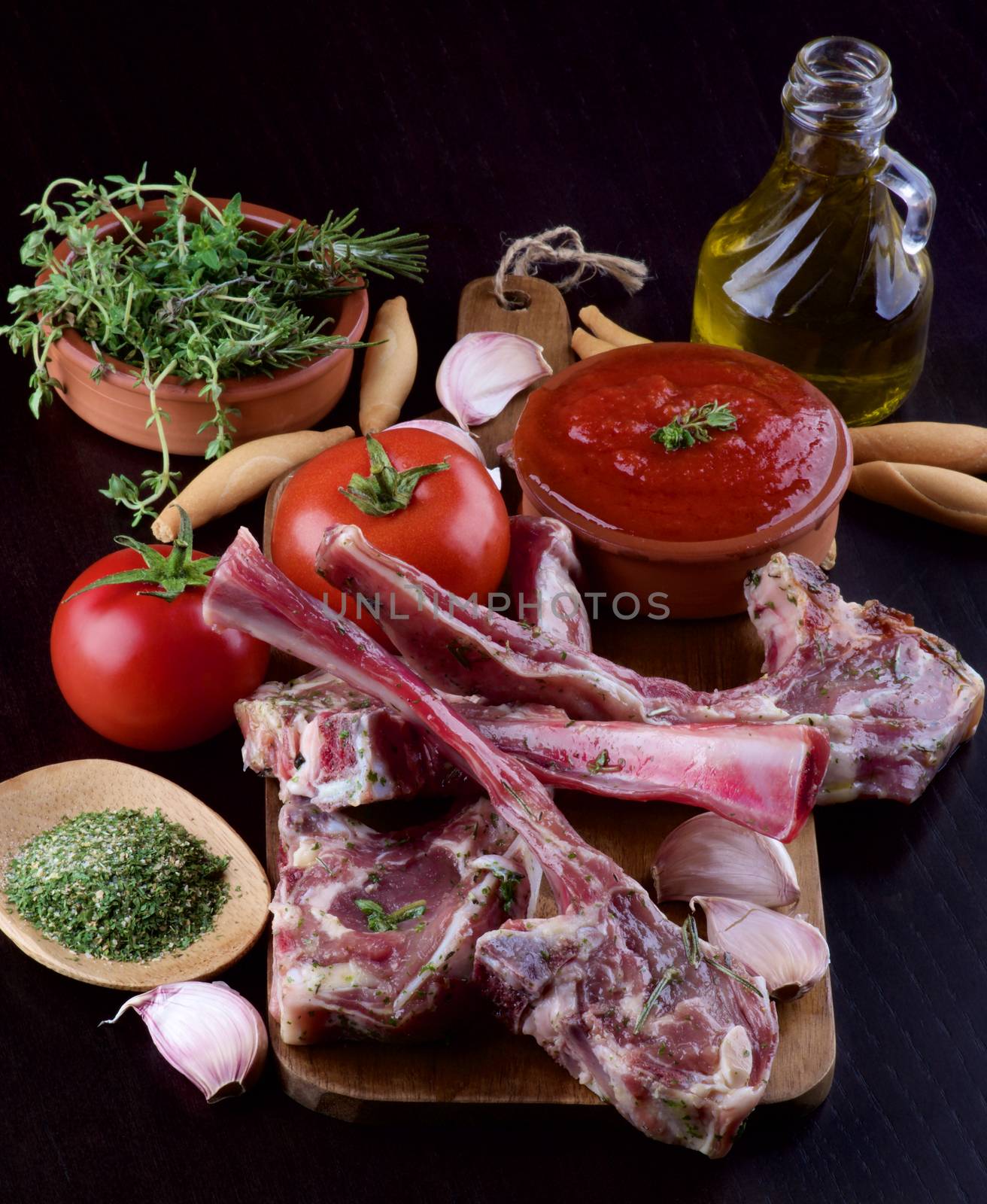 Ready to Roast Raw Lamb Ribs with Tomatoes, Sauce, Garlic, Herbs, Spices and Olive Oil on Wooden Cutting Board closeup on Dark Wooden background