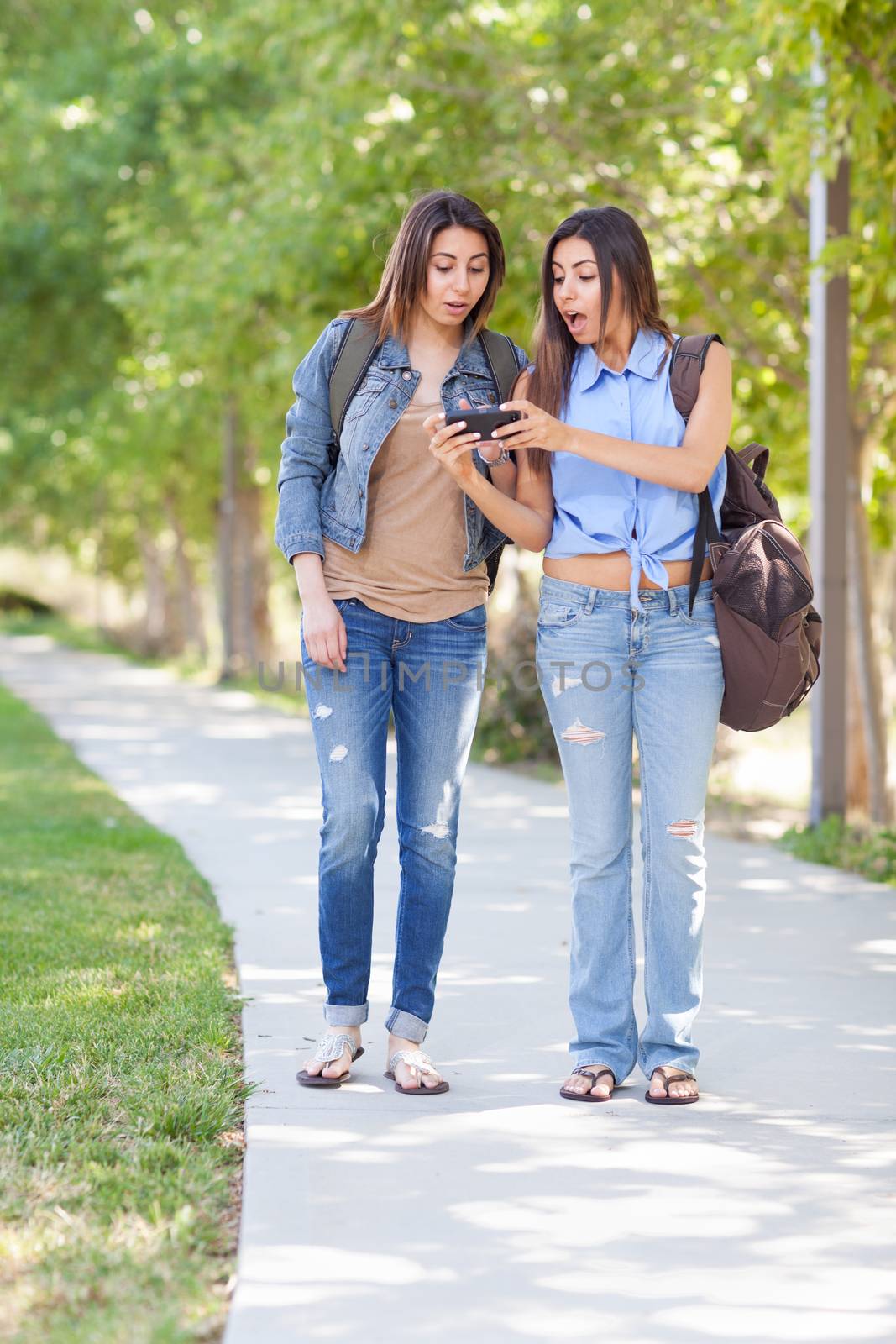 Two Beautiful Young Ethnic Twin Sisters With Backpacks Using A Smartphone Outside. by Feverpitched