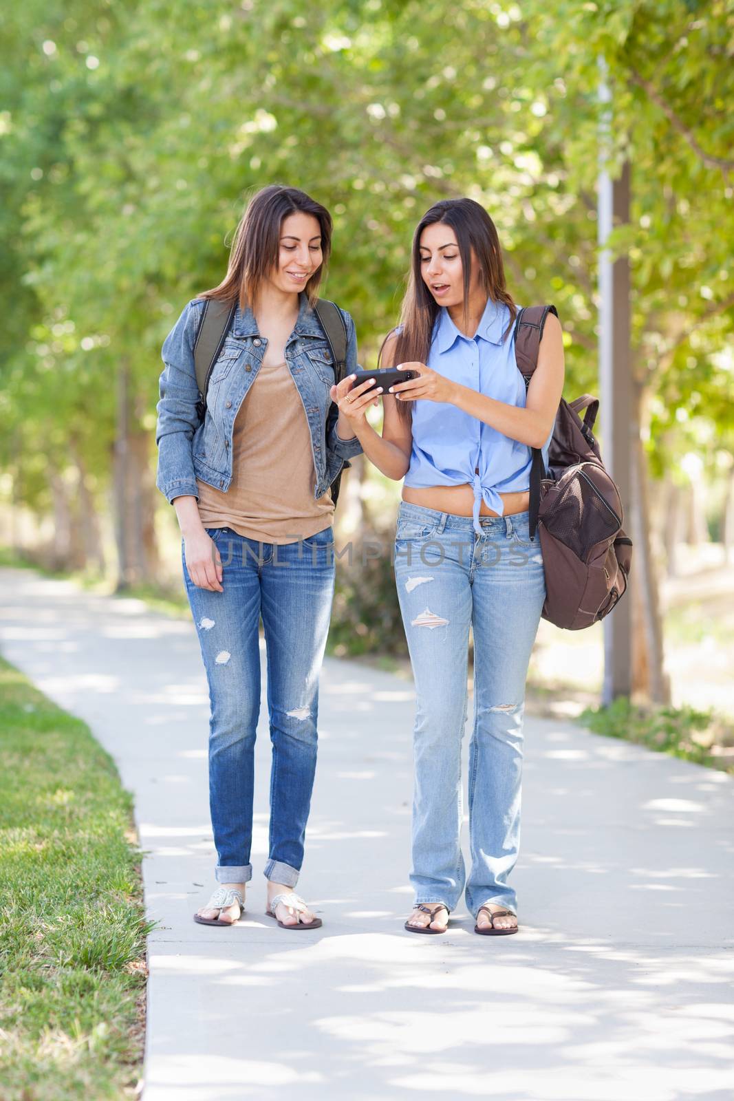 Two Beautiful Young Ethnic Twin Sisters With Backpacks Using A Smartphone Outside. by Feverpitched
