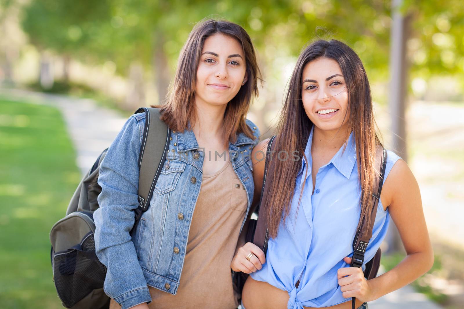 Two Beautiful Young Ethnic Twin Sisters With Backpacks Walking Outdoors. by Feverpitched