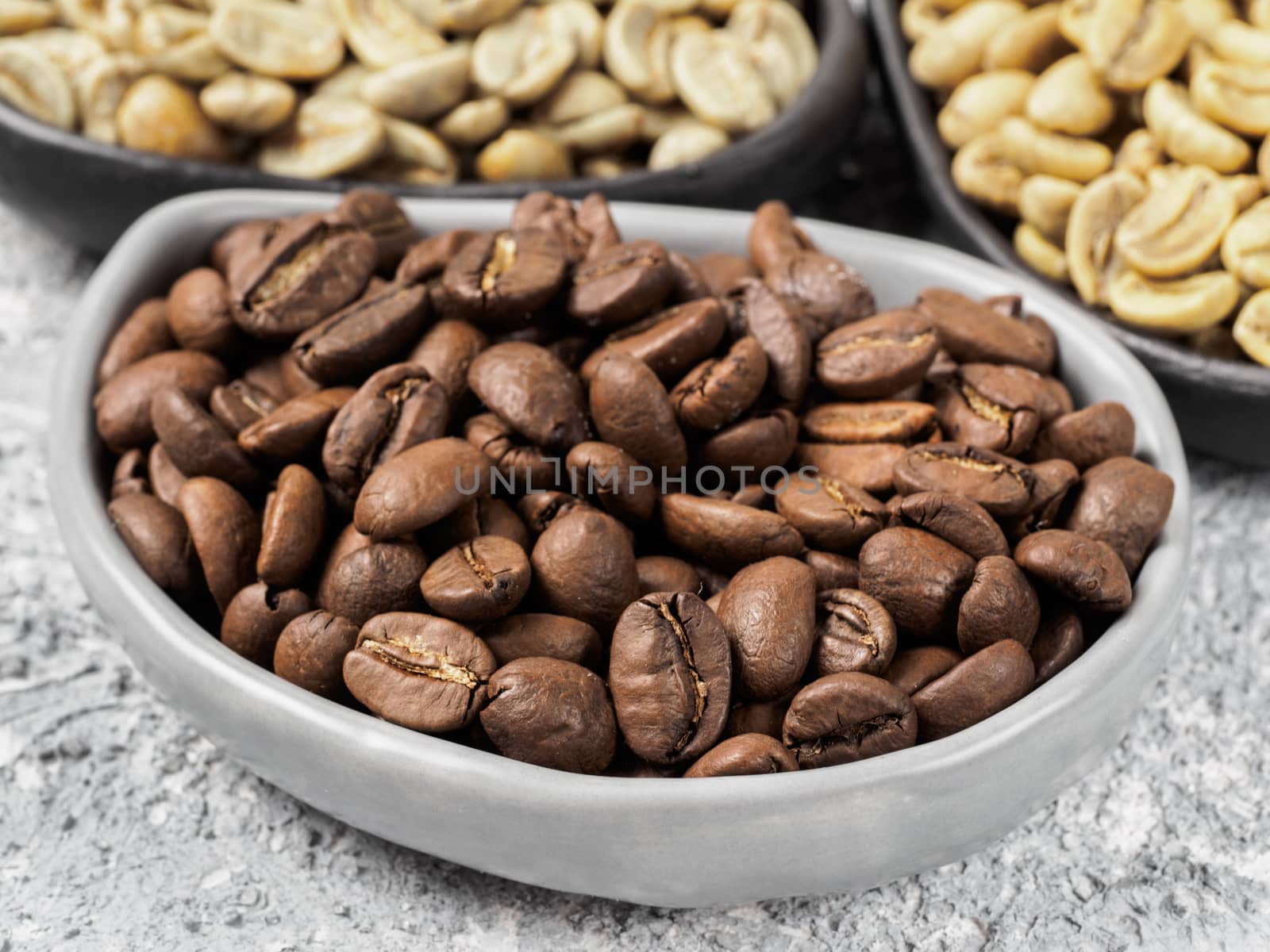 Close up view of roasted brown coffee beans in trendy plate on gray concrete background. Green and white raw coffee on background. Image with natural colors