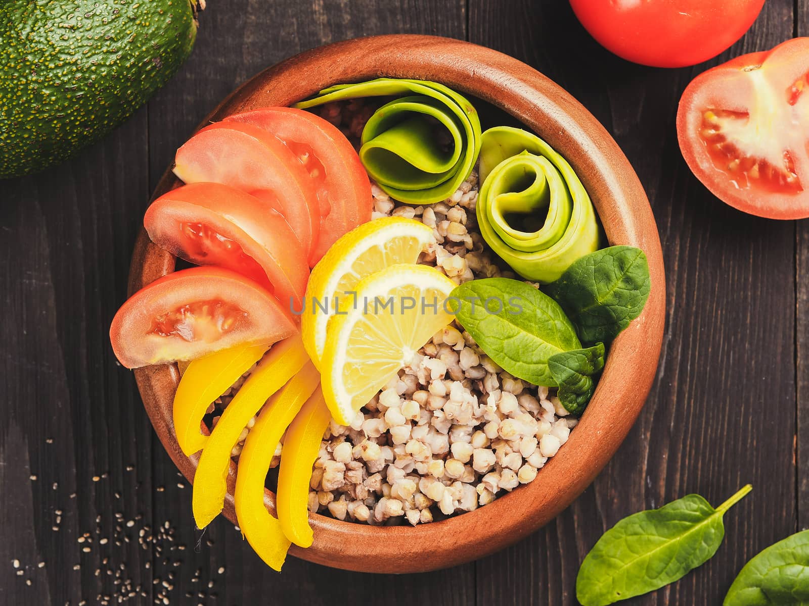 Top view of vegetarian buddha bowl with green buckwheat, spinach, avocado, tomatoes and yellow sweet pepper paprika on dark wooden table with copy space