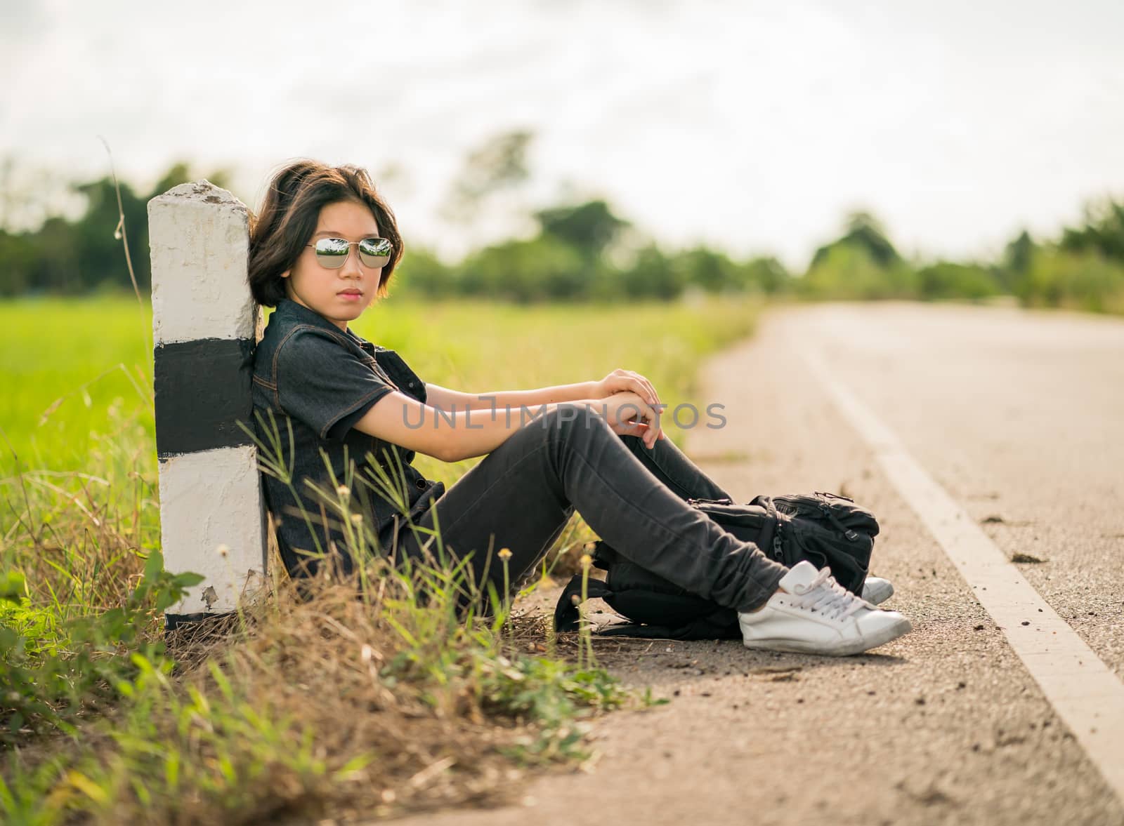 Young asian woman short hair and wearing sunglasses sit with backpack hitchhiking along a road wait for help in country road Thailand