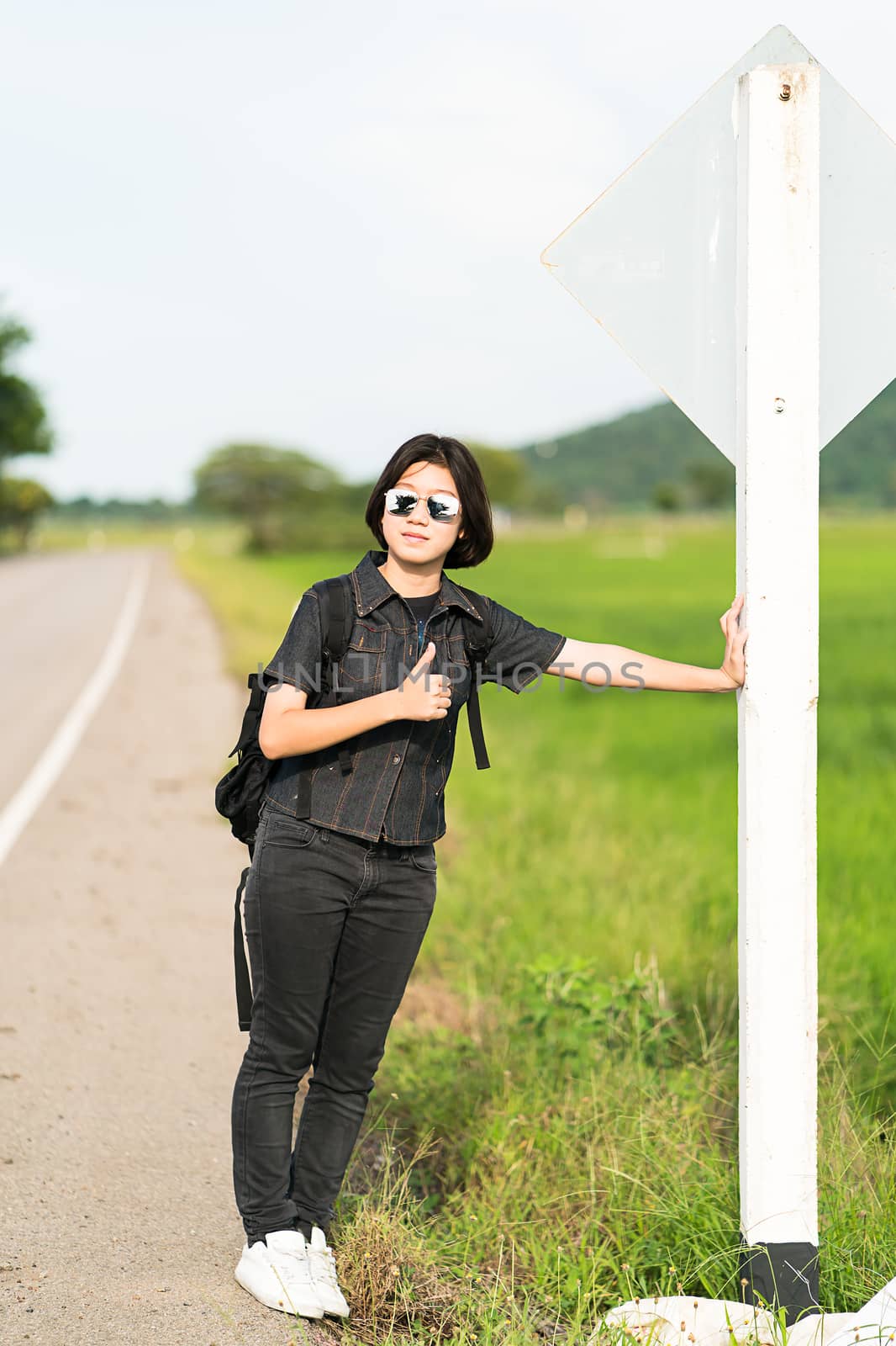 Young asian woman short hair and wearing sunglasses with backpack hitchhiking along a road in countryside Thailand