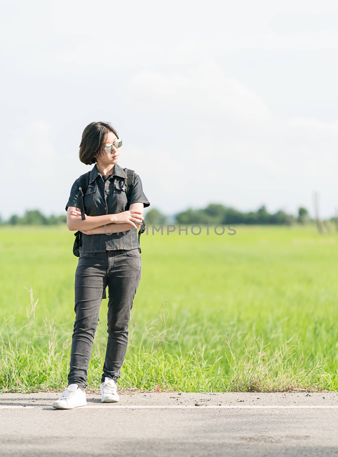 Young asian woman short hair and wearing sunglasses with backpack hitchhiking along a road in countryside Thailand