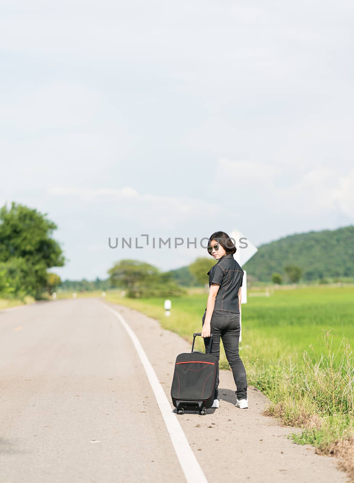 Young asian woman short hair and wearing sunglasses with luggage hitchhiking along a road in countryside Thailand