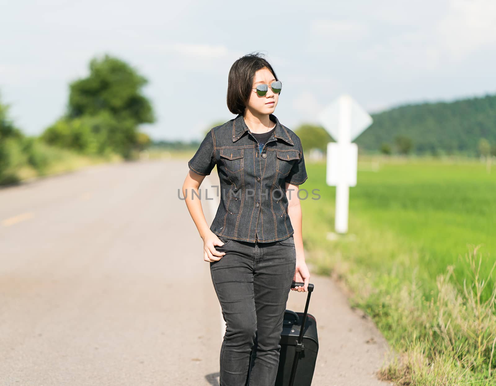 Young asian woman short hair and wearing sunglasses with luggage hitchhiking along a road in countryside Thailand