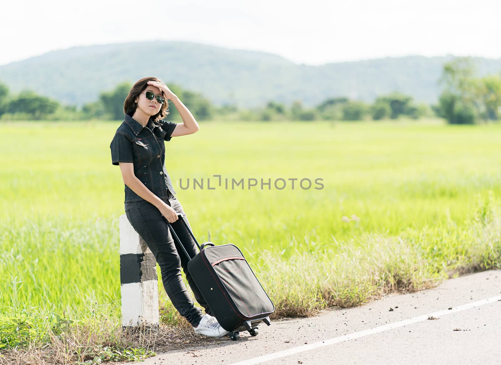 Young asian woman short hair and wearing sunglasses with luggage hitchhiking along a road in countryside Thailand