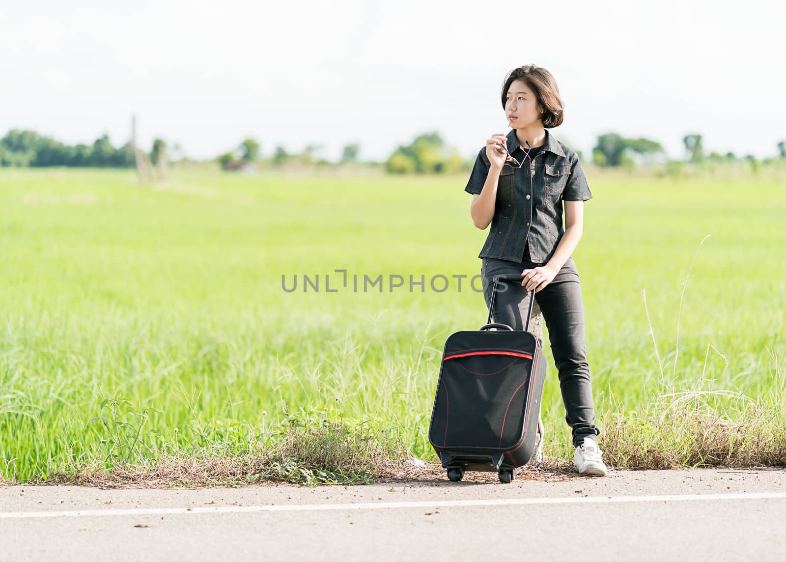 Young asian woman short hair and wearing sunglasses with luggage hitchhiking along a road in countryside Thailand