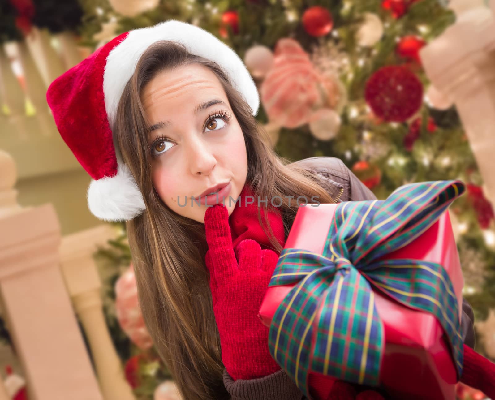 Thinking Girl Wearing A Christmas Santa Hat with Bow Wrapped Gift In Front of Decorated Tree. by Feverpitched