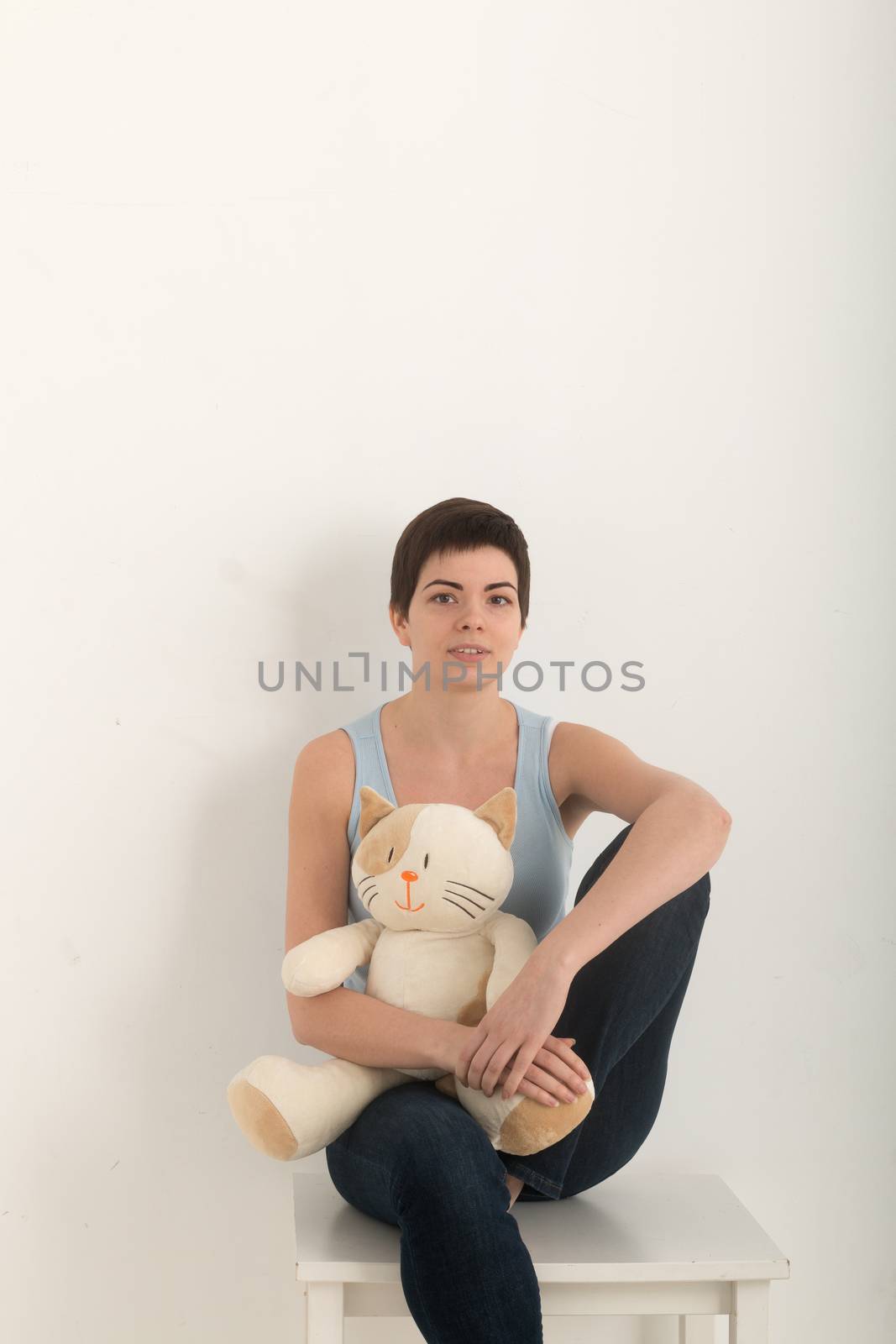Vertical image of young girl looking at camera, portrait of a beautiful brunette woman with a lovely plush toy cat in arms, she sits on a white table on white wall background. Shot with flare. by MSharova