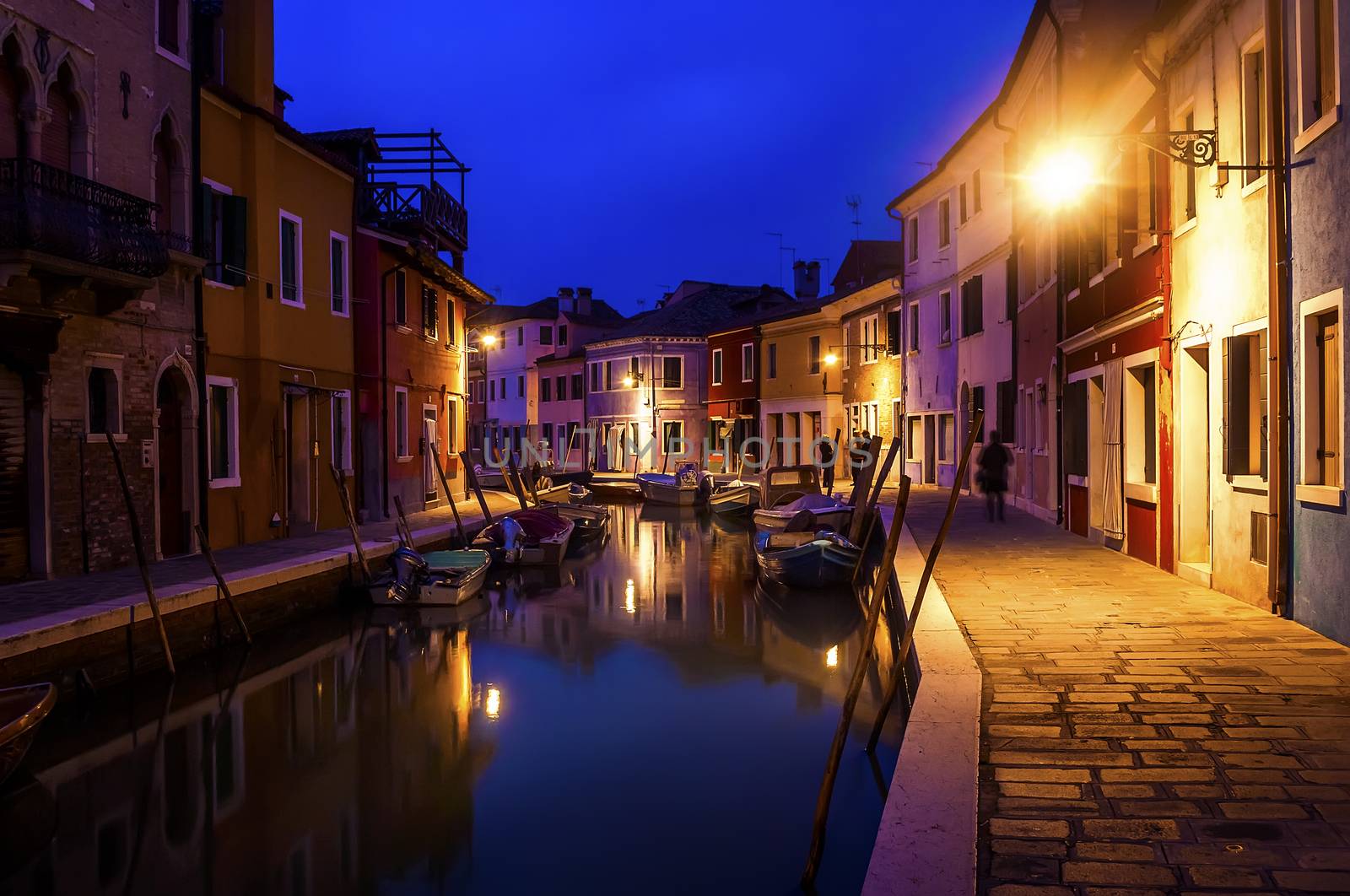 Beautiful evening cityscape of Burano island in Venice. Colored buildings are illuminated by street lights reflected in a water canal. Boats are parked there.