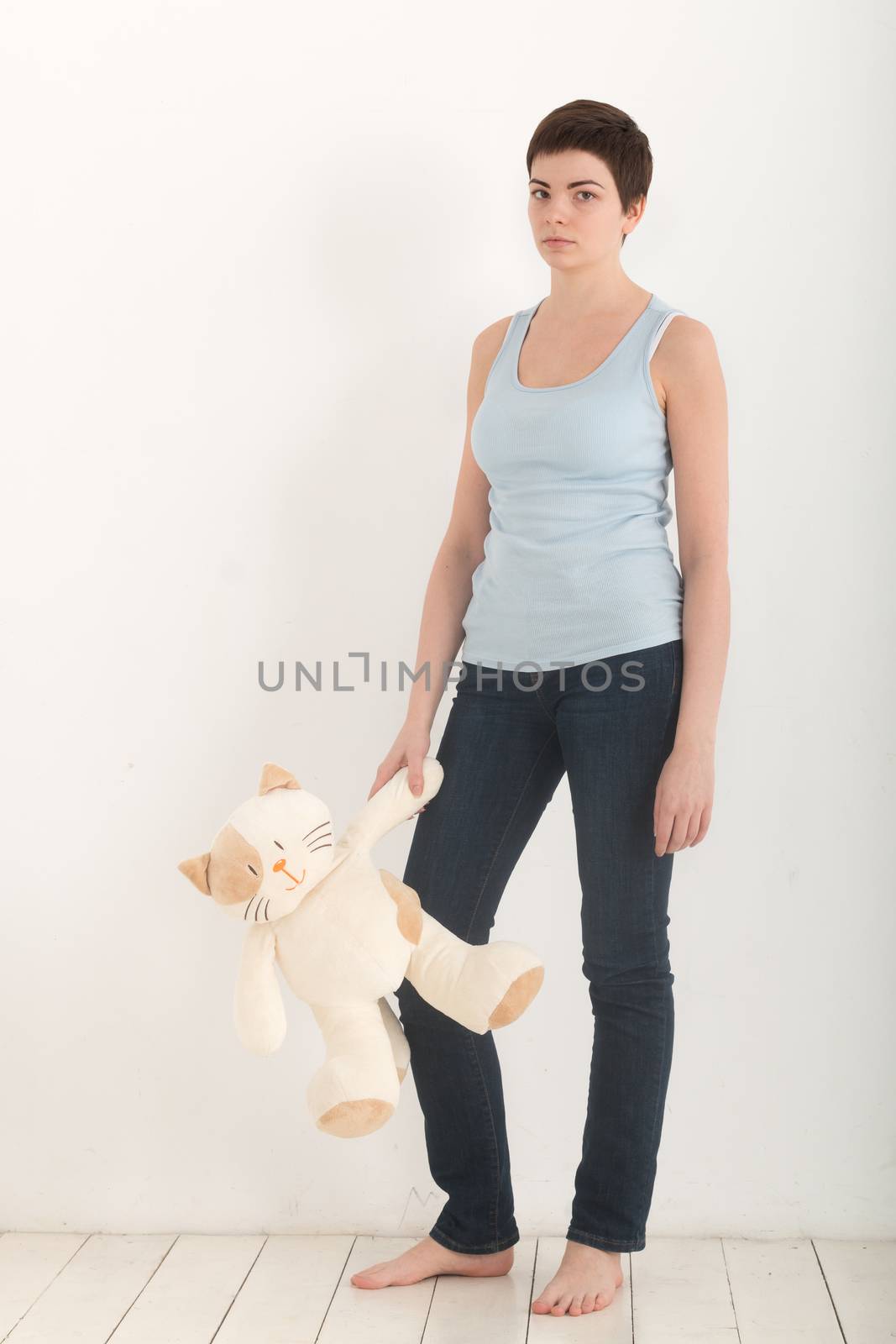 Pretty girl standing over white background barefoot with plush toy cat in her arm, looking seriously and upset. Full length of a brunette woman with short hair in a blue t-shirt and denim jeans