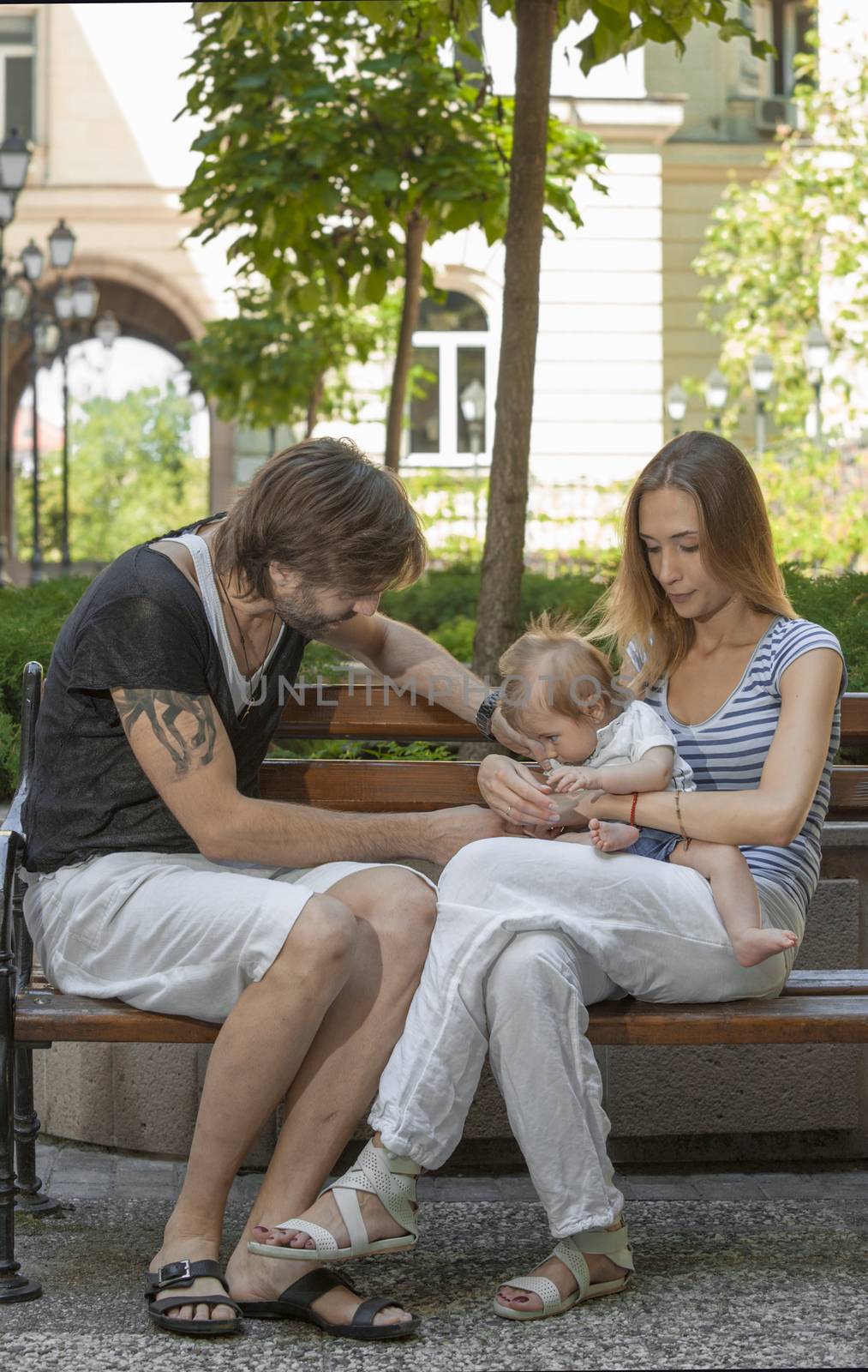 Happy young beautiful family of three is sitting on a bench outdoors in a european city.