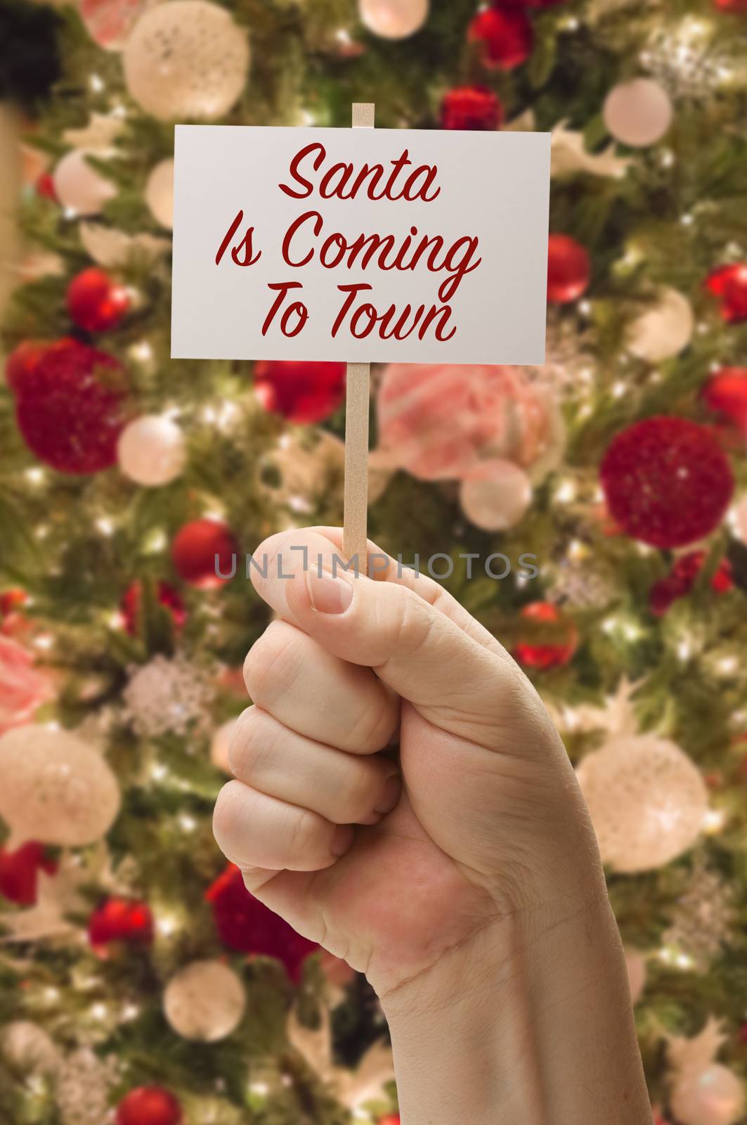 Hand Holding Santa Is Coming To Town Card In Front of Decorated Christmas Tree.