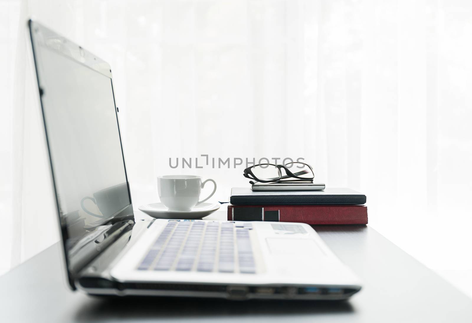 Close up laptop placed on desktop with coffee cup and glasses on book in room at home office