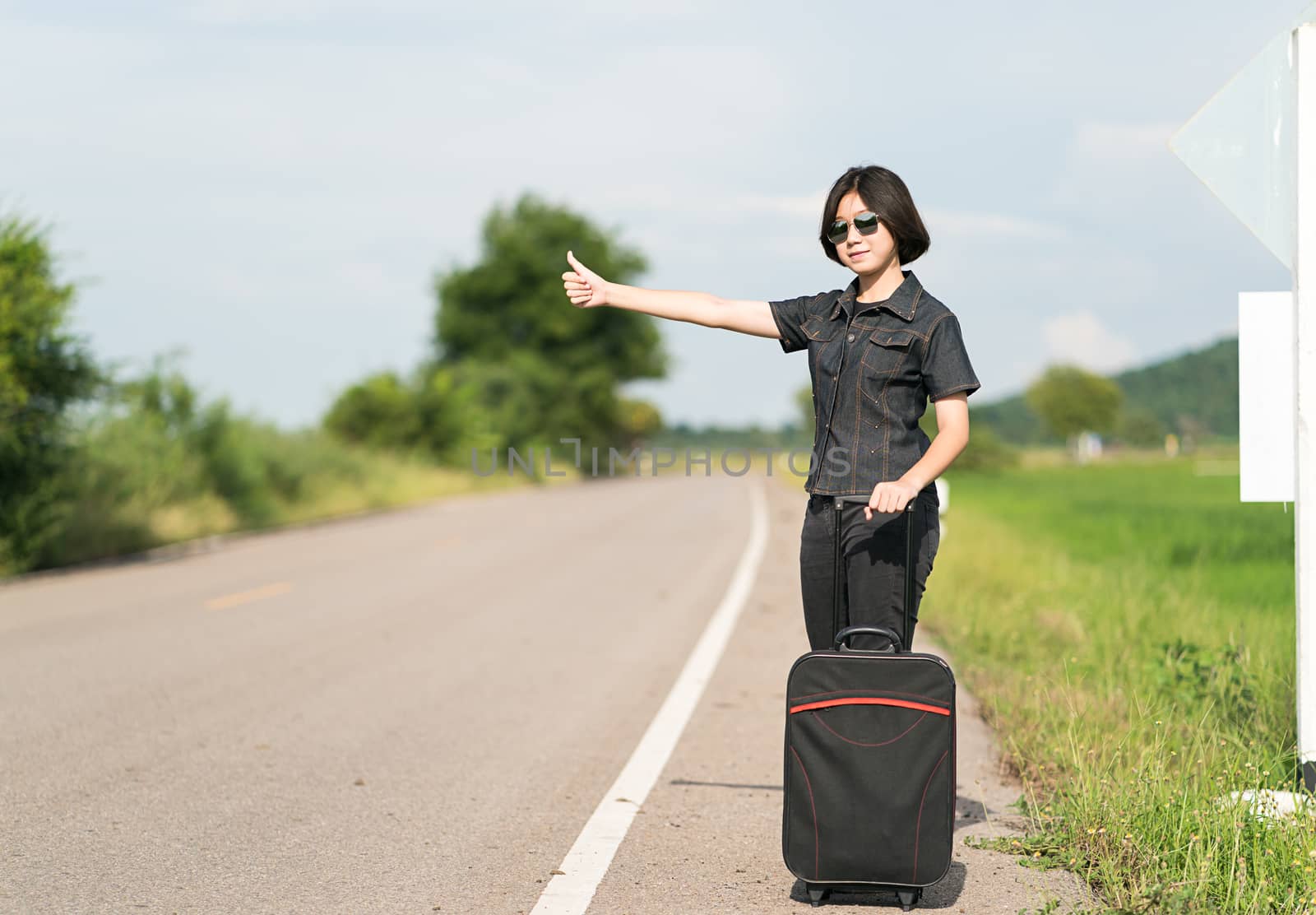 Young asian woman short hair and wearing sunglasses with luggage hitchhiking along a road and thumbs up in country road Thailand